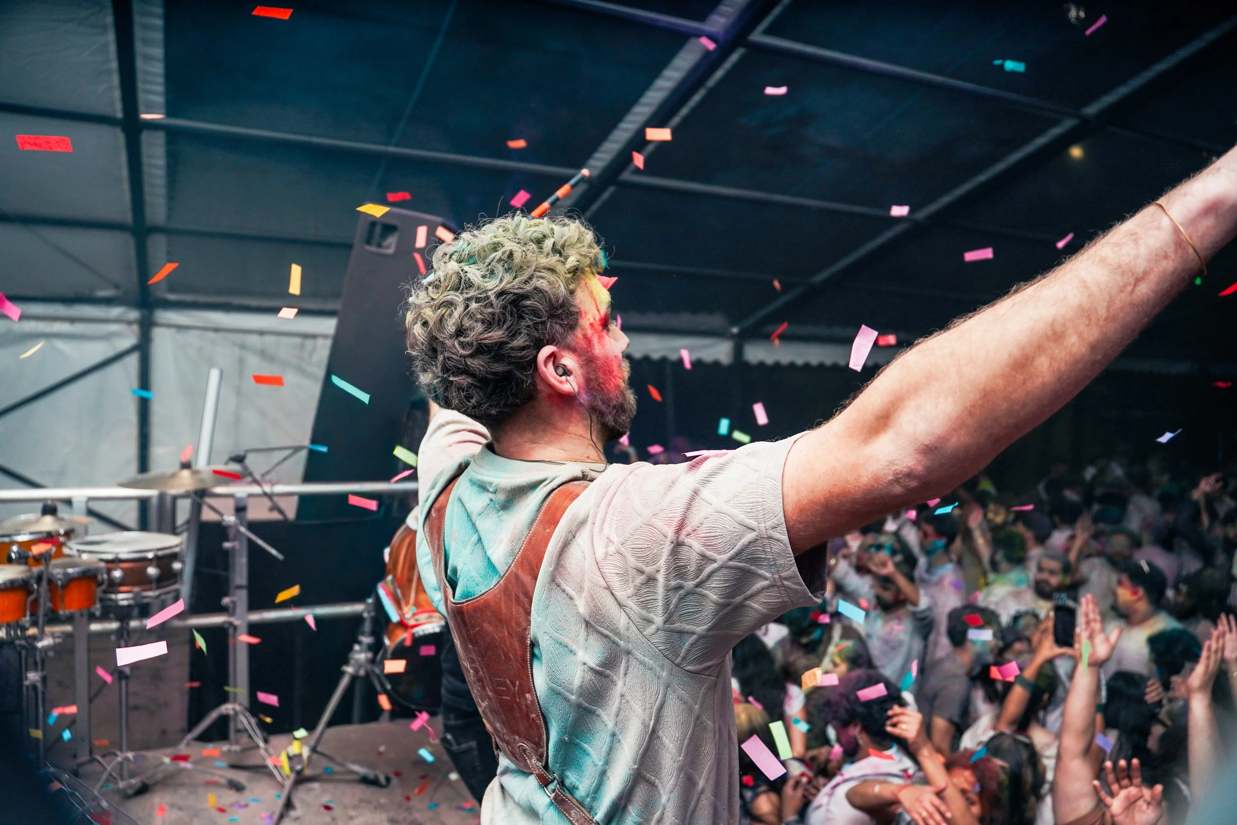 A man with curly hair and a beard, wearing a beige top, is on a stage with confetti falling around him. He is facing a crowd with his arms outstretched, celebrating at a lively concert or event.