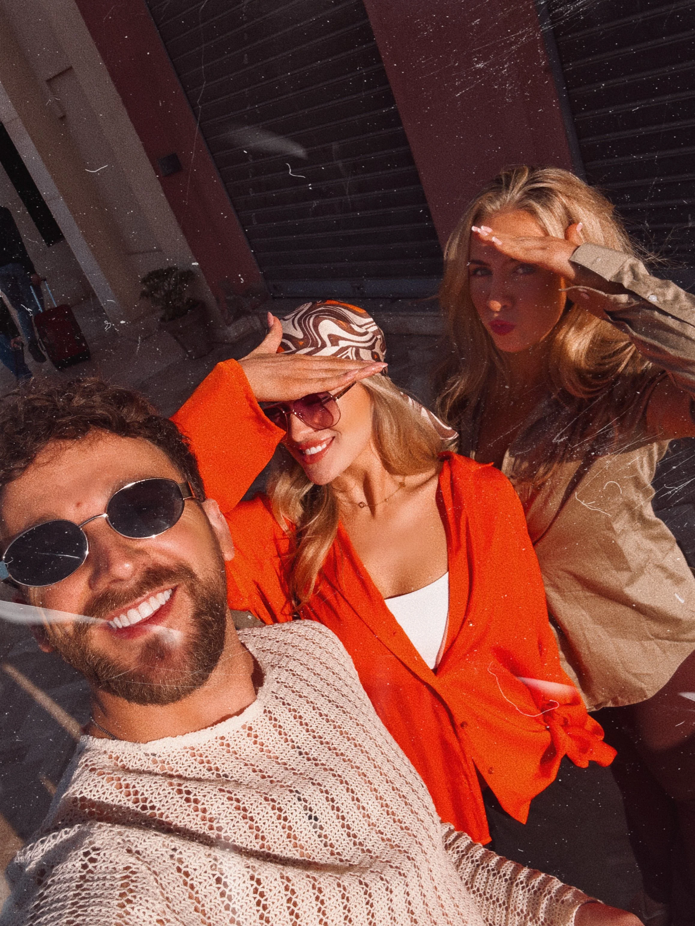 A group of three friends taking a selfie outdoors, shielding their eyes from the sunlight with their hands. The man in the foreground is smiling and wearing sunglasses and a knitted sweater. The woman in the middle is wearing sunglasses, a patterned headscarf, and an orange shirt. The woman on the right is also shielding her eyes from the sun with her hand, wearing a beige jacket. They are standing in front of a dark shutter and a building with a potted plant and luggage in the background.