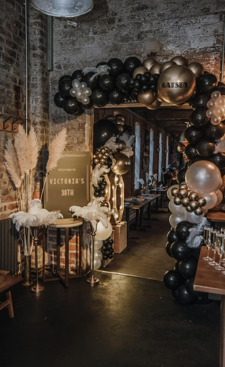 Decorative entrance with black, gold, and white balloons arching over a hallway, feathers, pampas grass, a sign that reads 'Welcome to Victoria's 30th,' and a brick wall background. Tobi Ripley. Roaming bongo drummer. Percussionist. event drummer. we