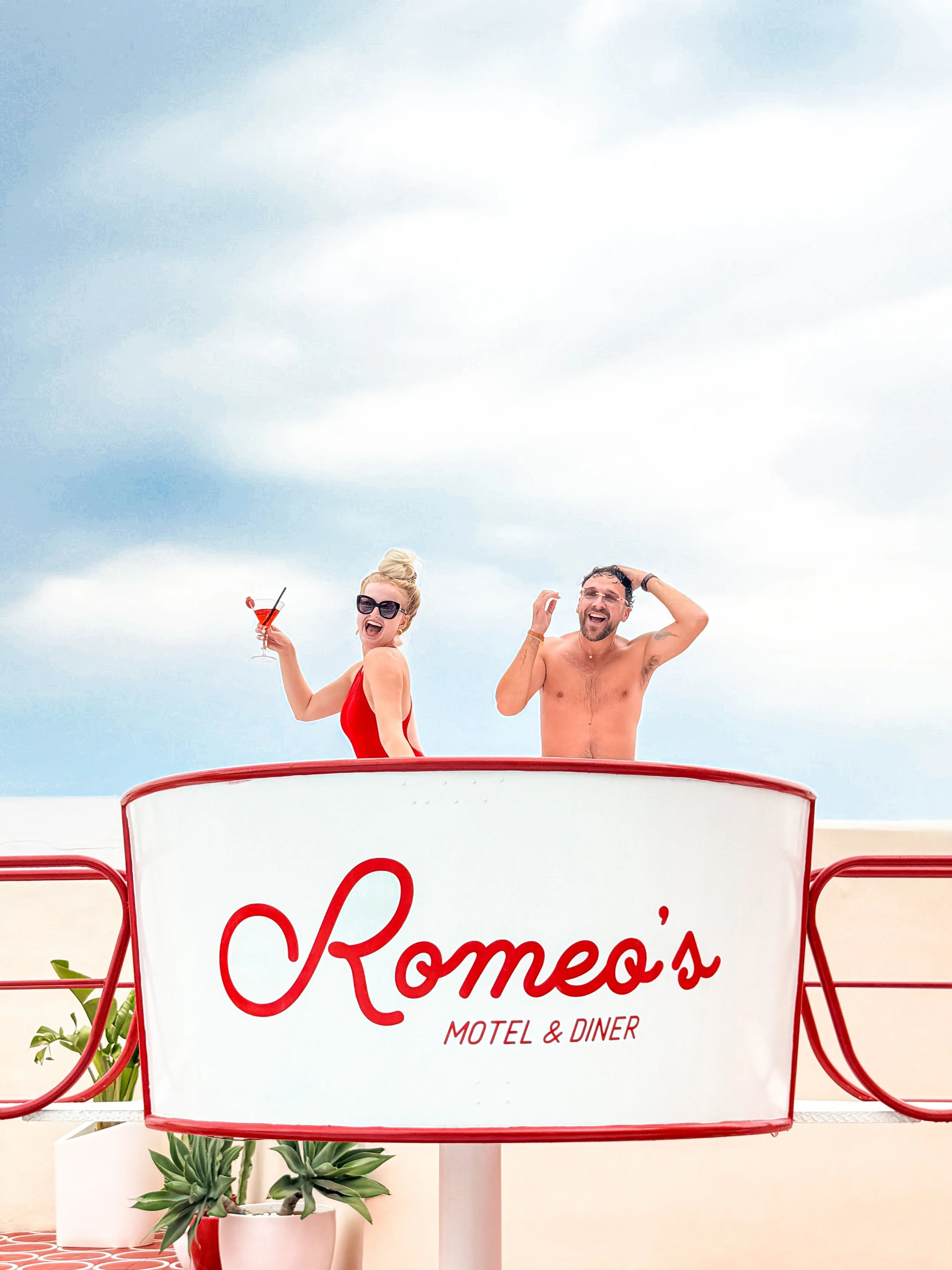 Two people, a woman in a red swimsuit and a man without a shirt, are on a boat or elevated platform at a motel and diner called Romeo's. The woman holds a drink and sunglasses, both are smiling and appear to be enjoying themselves. The sky in the background is partly cloudy.
