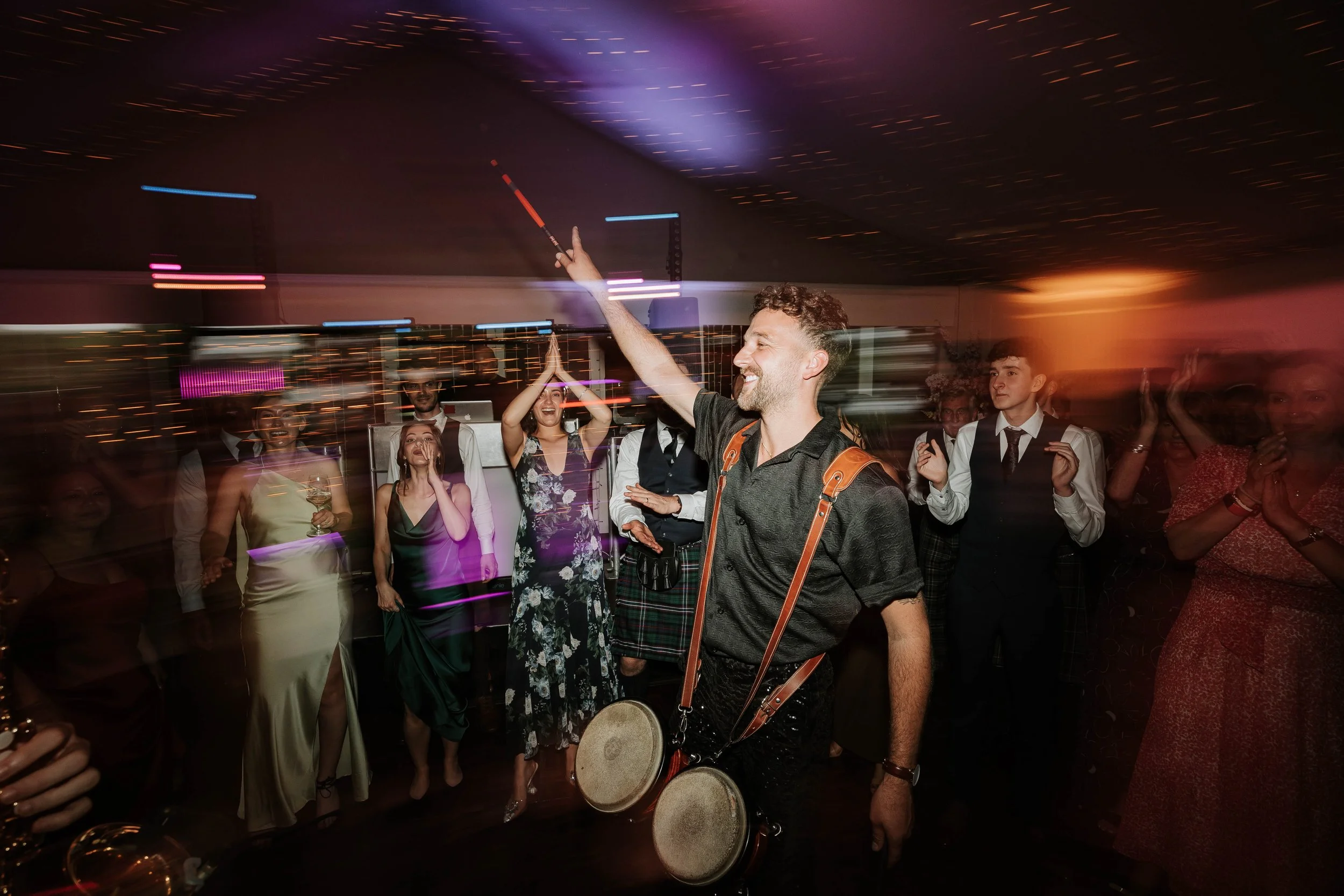 A man with drums around his neck dances at a party or celebration with a group of people watching and clapping in the background, under colorful lights.