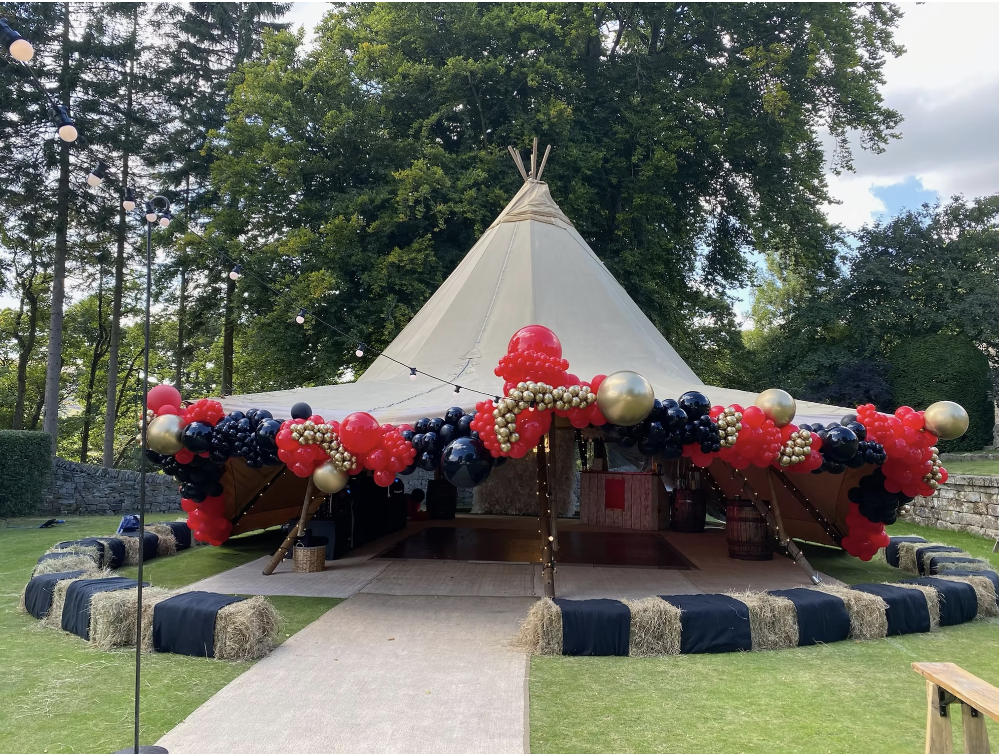Decorated party tent with red, black, gold, and silver balloons, set outdoors on a grassy area with string lights and trees in the background. Tobi Ripley. Roaming bongo drummer. Percussionist. event drummer. wedding entertainment. festival drummer.