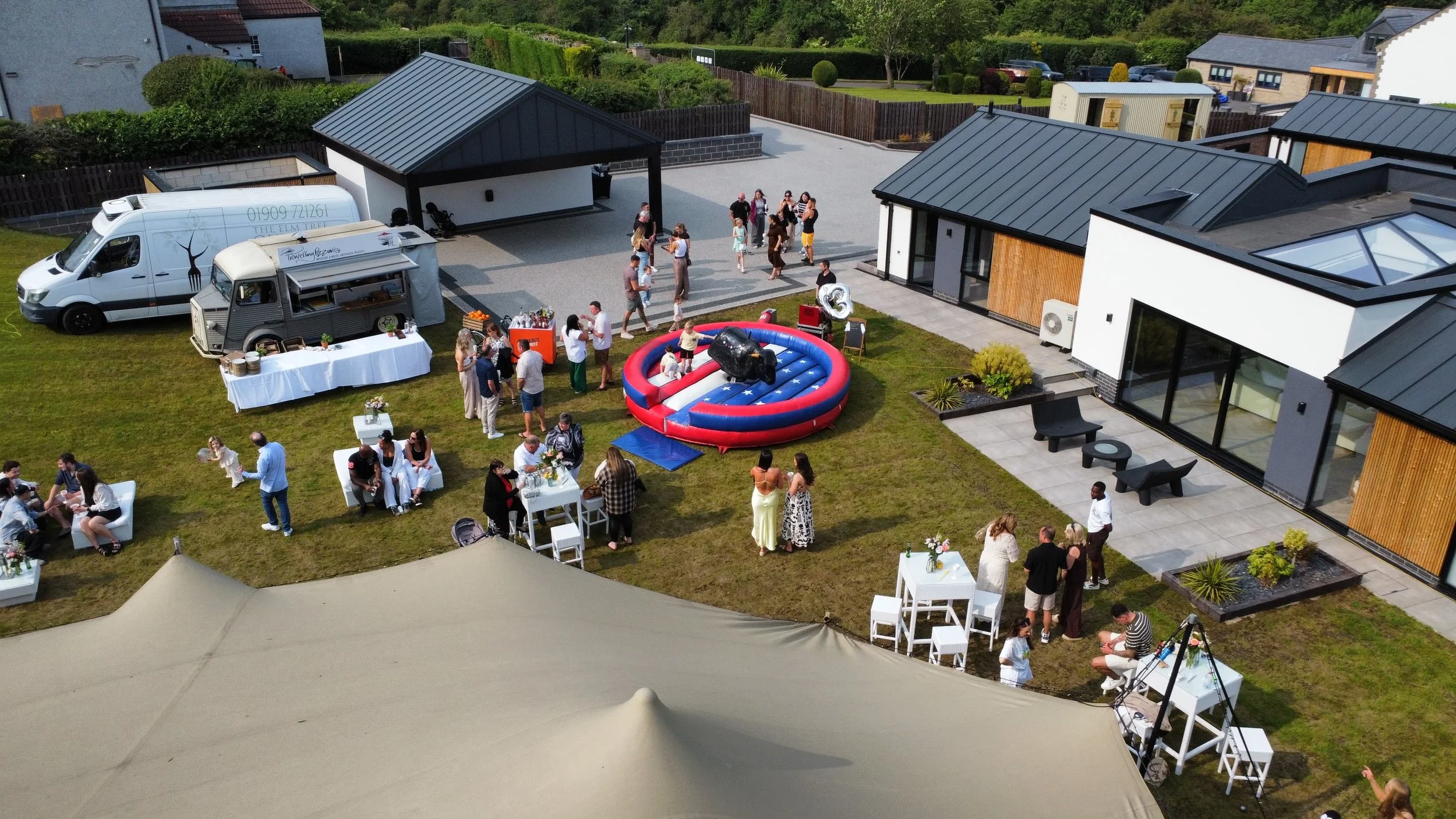 A social gathering in a backyard with groups of people chatting, some sitting on white chairs, and others standing around. There's an inflatable rodeo bull in the center, food and drink stands, and tables with flowers. The area is surrounded by modern houses with large windows, a patio with outdoor seating, and a green lawn.