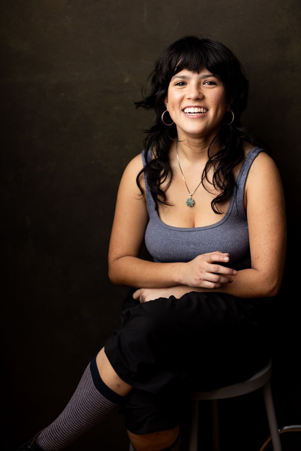 A woman with black, wavy hair, wearing a gray tank top, earrings, and a necklace, seated on a stool against a dark background, smiling at the camera.