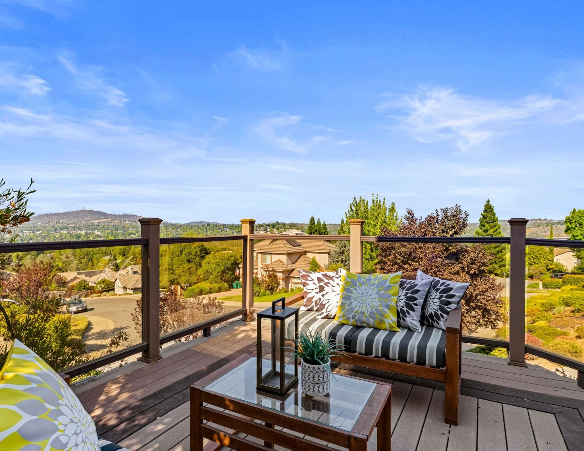 A backyard patio with a wooden seating area featuring black and white floral pillows, a small table with a lantern and potted plant, overlooking a neighborhood with houses, trees, and a hill under a blue sky.