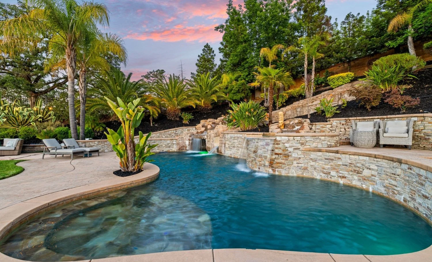 Luxury backyard with resort-style swimming pool, stone waterfall features, tropical landscaping, and lounge seating at sunset in an upscale California home.