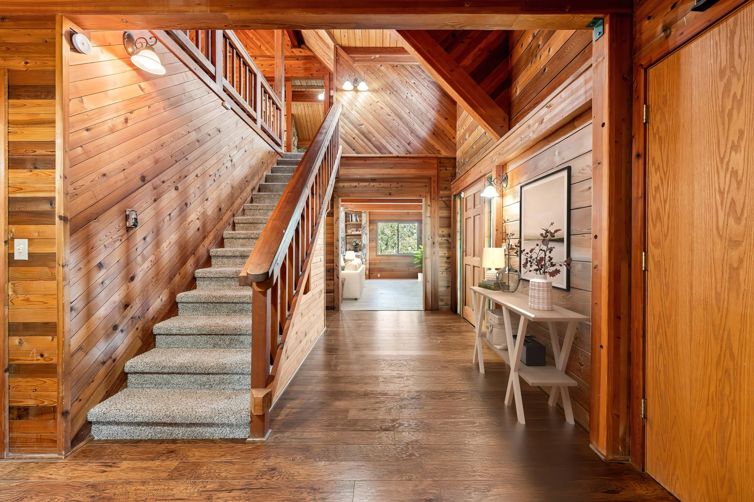 Interior view of a wooden house hallway with stairs, a small console table, lamps, framed artwork, and natural light from a window at the end.
