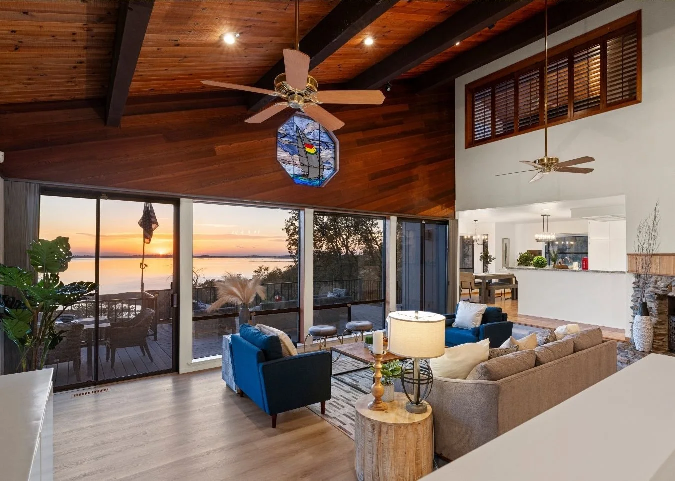 Living room with large floor-to-ceiling windows overlooking a water view at sunset, wooden ceiling with ceiling fans, modern furniture, and a sliding glass door leading to an outdoor deck.