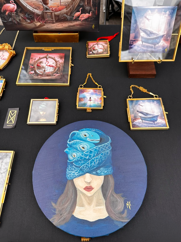 MADX Union Square Holiday Market NYC booth display featuring a hand-painted oil original of Justice of the Deep on round canvas, tiny gold-framed art prints and ornaments including Flamingo, Girl on Swing and Magic Whale Tub by NYC artist MADX.