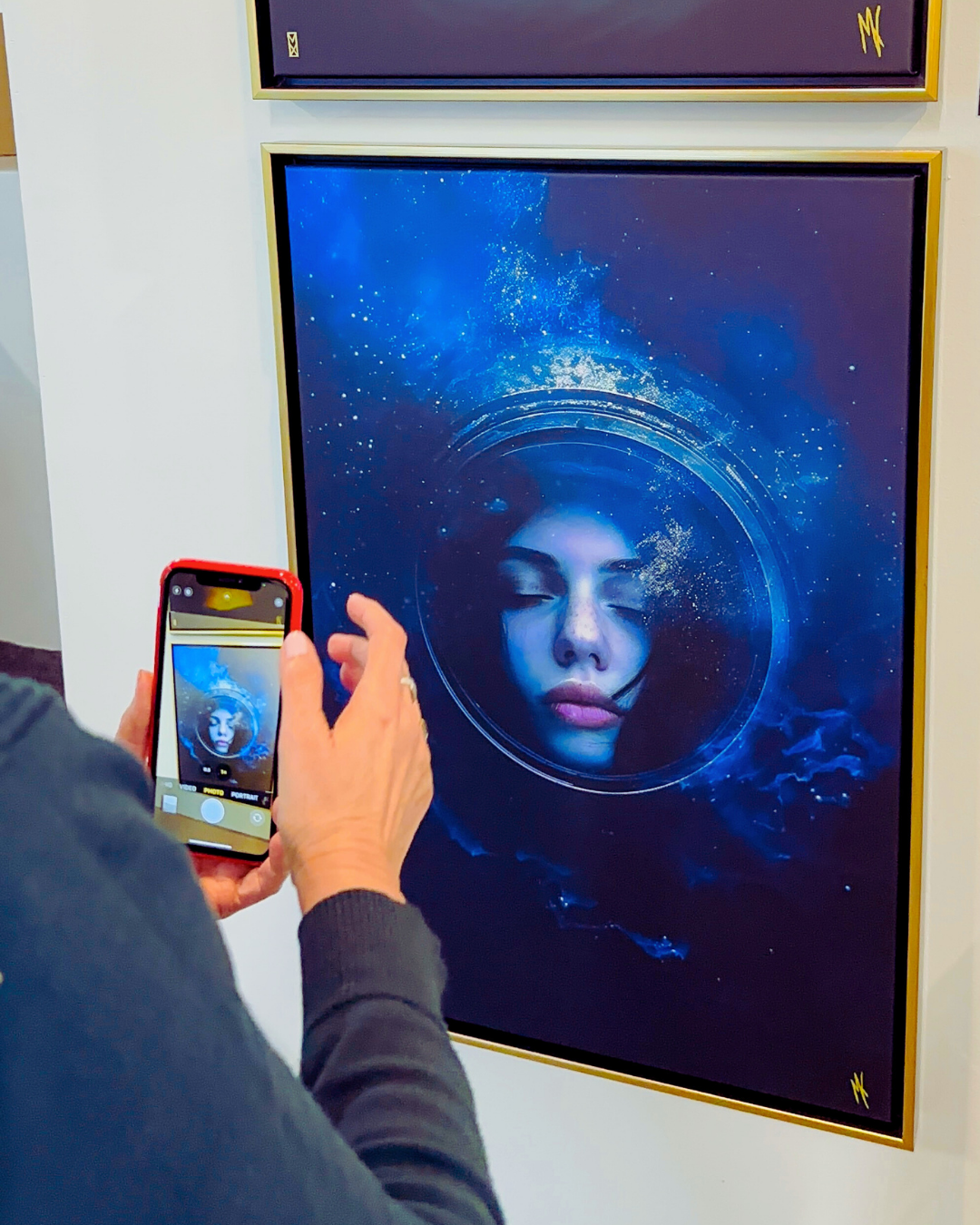 Gallery visitor photographing MADX Serenity surreal fine art canvas print in gold float frame at Art Expo New York 2025. Deep blue mixed-media artwork of ethereal female face by NYC artist MADX.