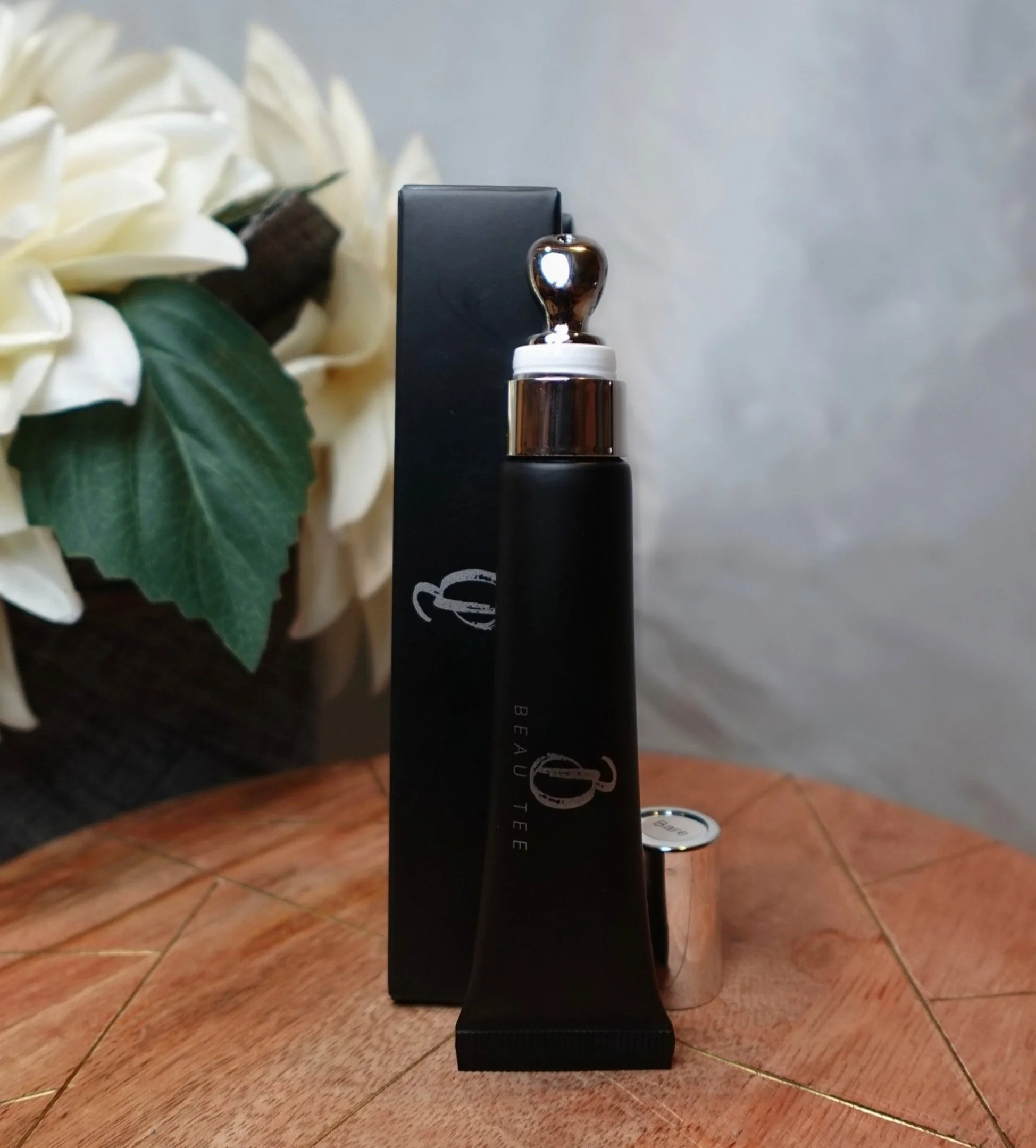 Beau Tee Luxe Gloss in a black tube with a silver applicator displayed beside its packaging.