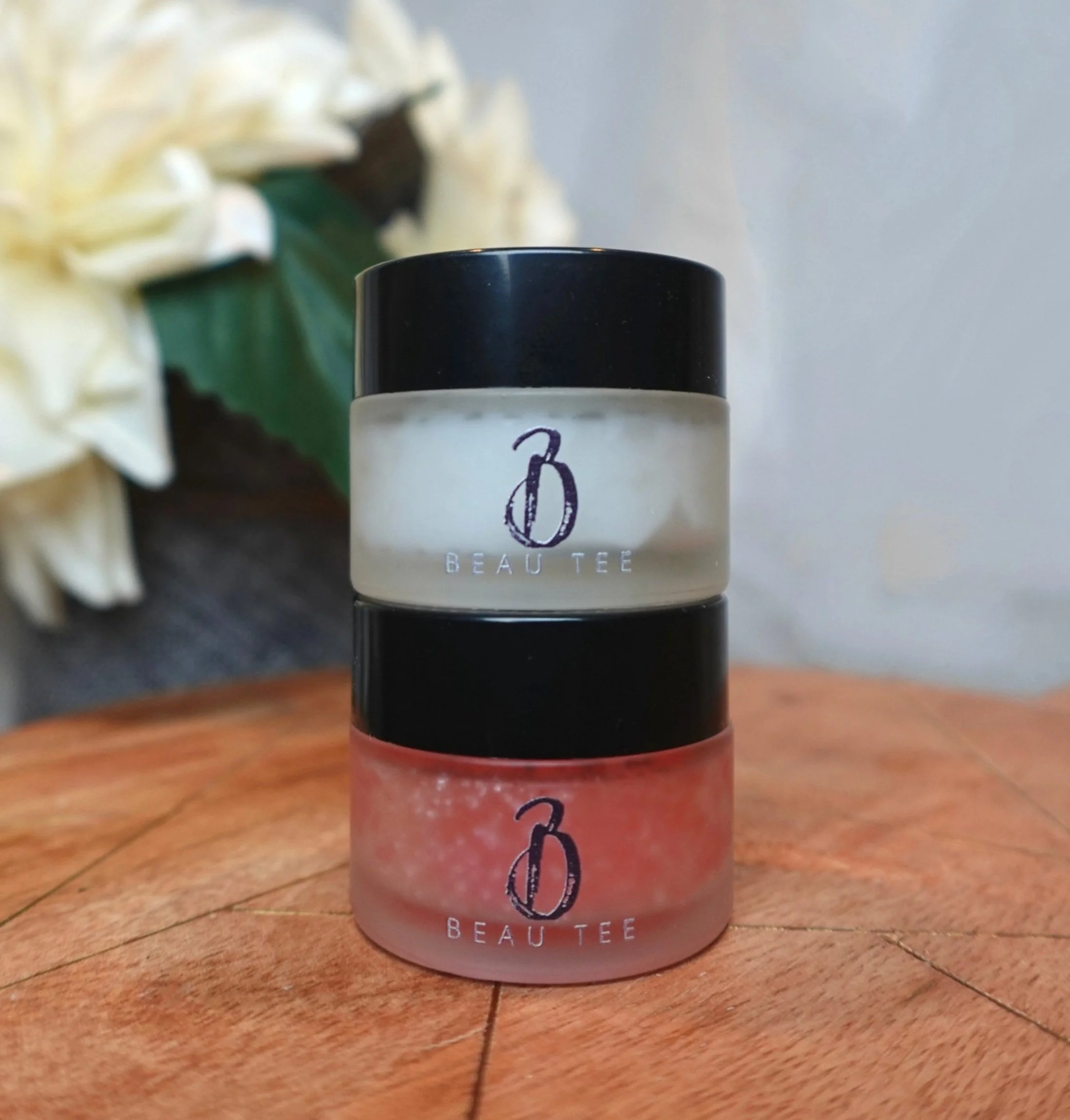 Beau Tee Luxe Lip Exfoliator with pink scrub in a clear jar displayed on a wooden surface.