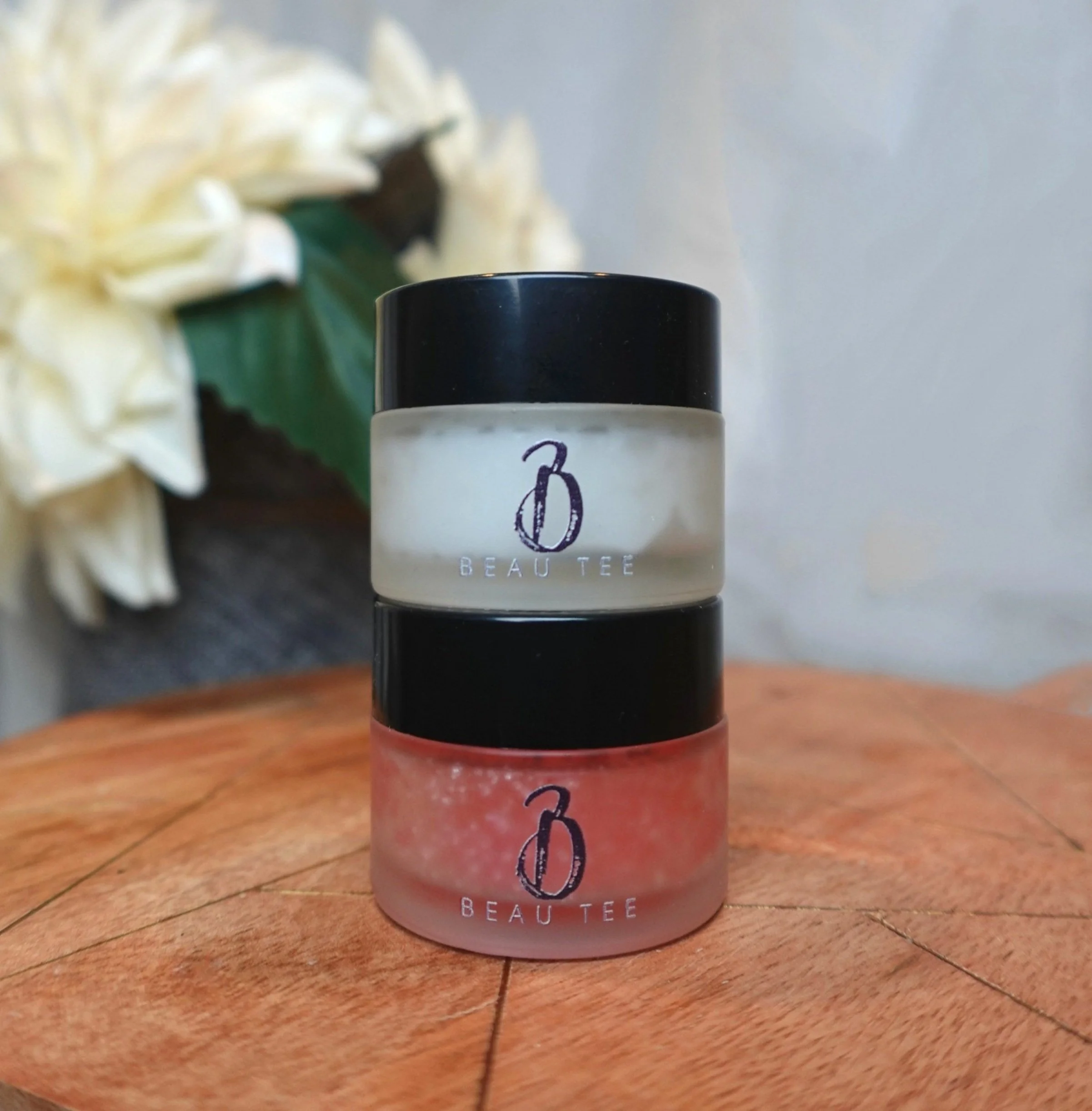 Beau Tee Luxe Lip Exfoliator with pink scrub in a clear jar displayed on a wooden surface.