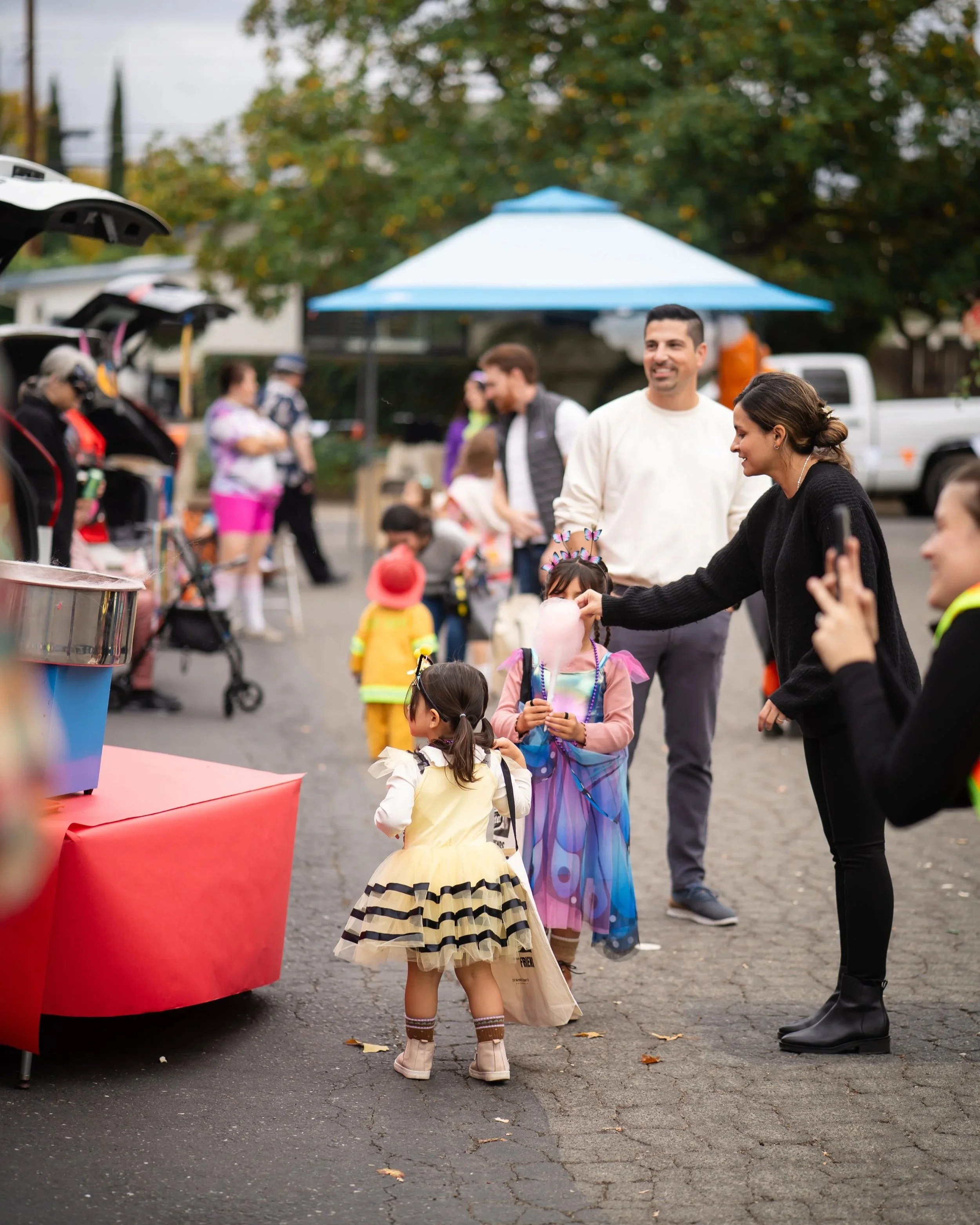 People, including children dressed in costumes, gather at an outdoor event on a city street with trees and parked cars in the background.