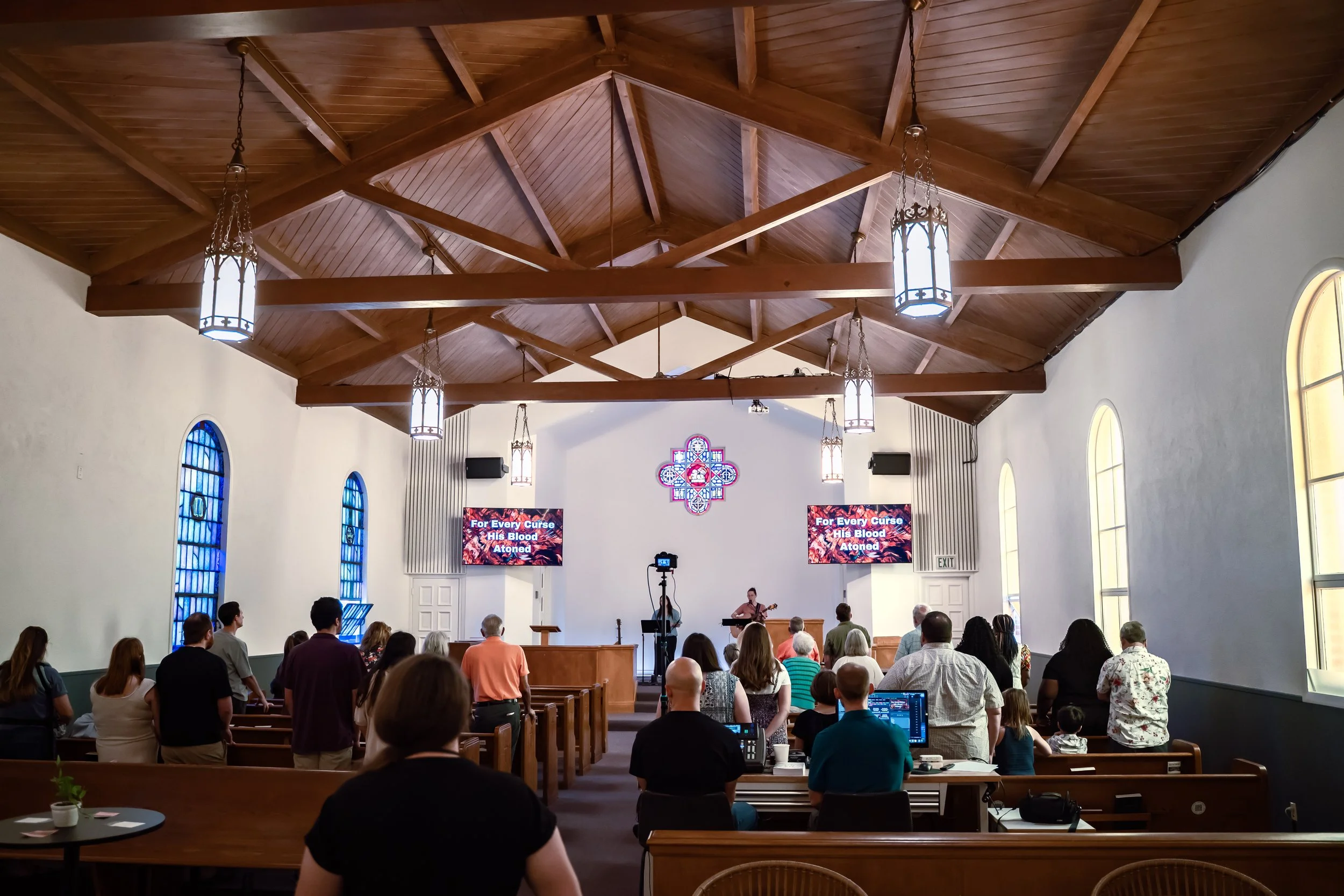 A welcoming church in Sacramento, CA. Interior of a church with a wooden ceiling, stained glass windows, and a congregation listening to a speaker and musician at the front, with screens displaying the choir lyrics.