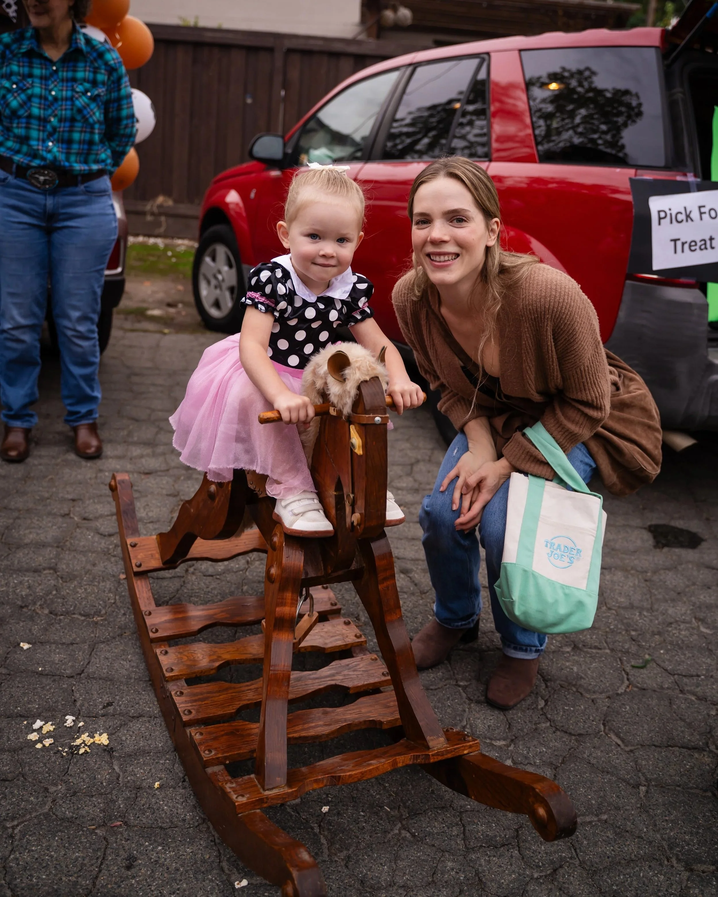 A young girl riding a wooden rocking horse with an adult woman kneeling beside her, smiling. In the background, a red car with a sign that reads 'Pick for Treats' and balloons tied to a fence.