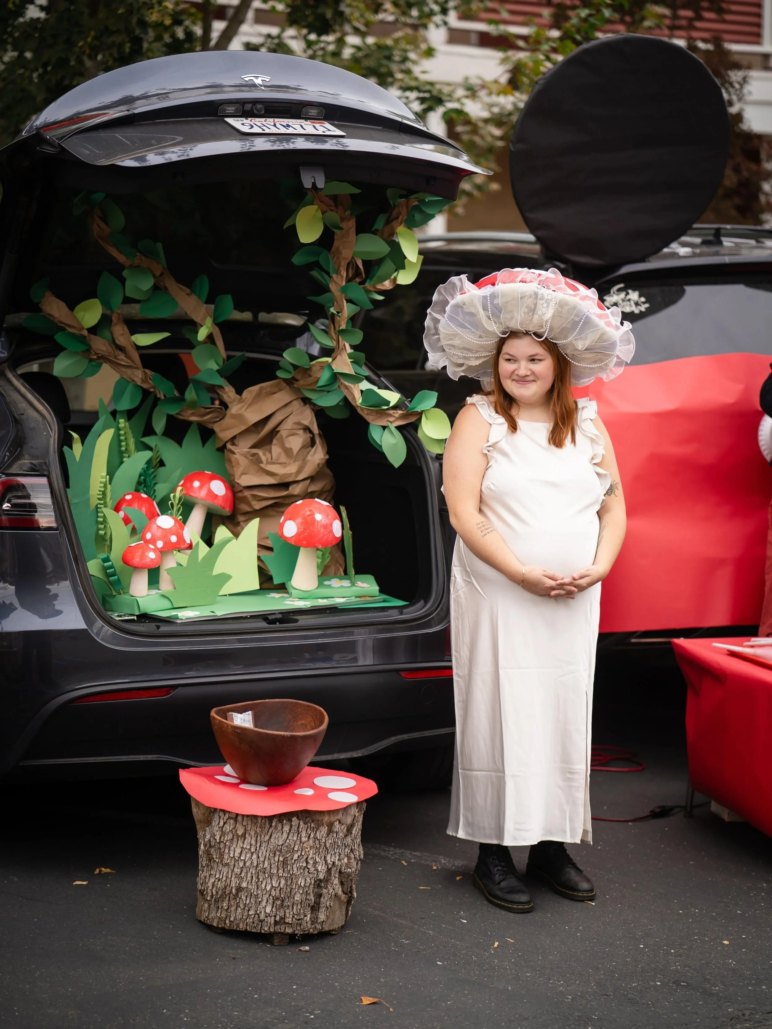 A woman dressed in a white gown and a large, decorative hat with pink and white ruffles stands next to a decorated car trunk. The trunk is decorated with paper and fabric trees, mushrooms, and plants, creating a whimsical forest scene. There is a tree stump with a bowl on top in front of the woman. The background shows part of a residential building and trees.