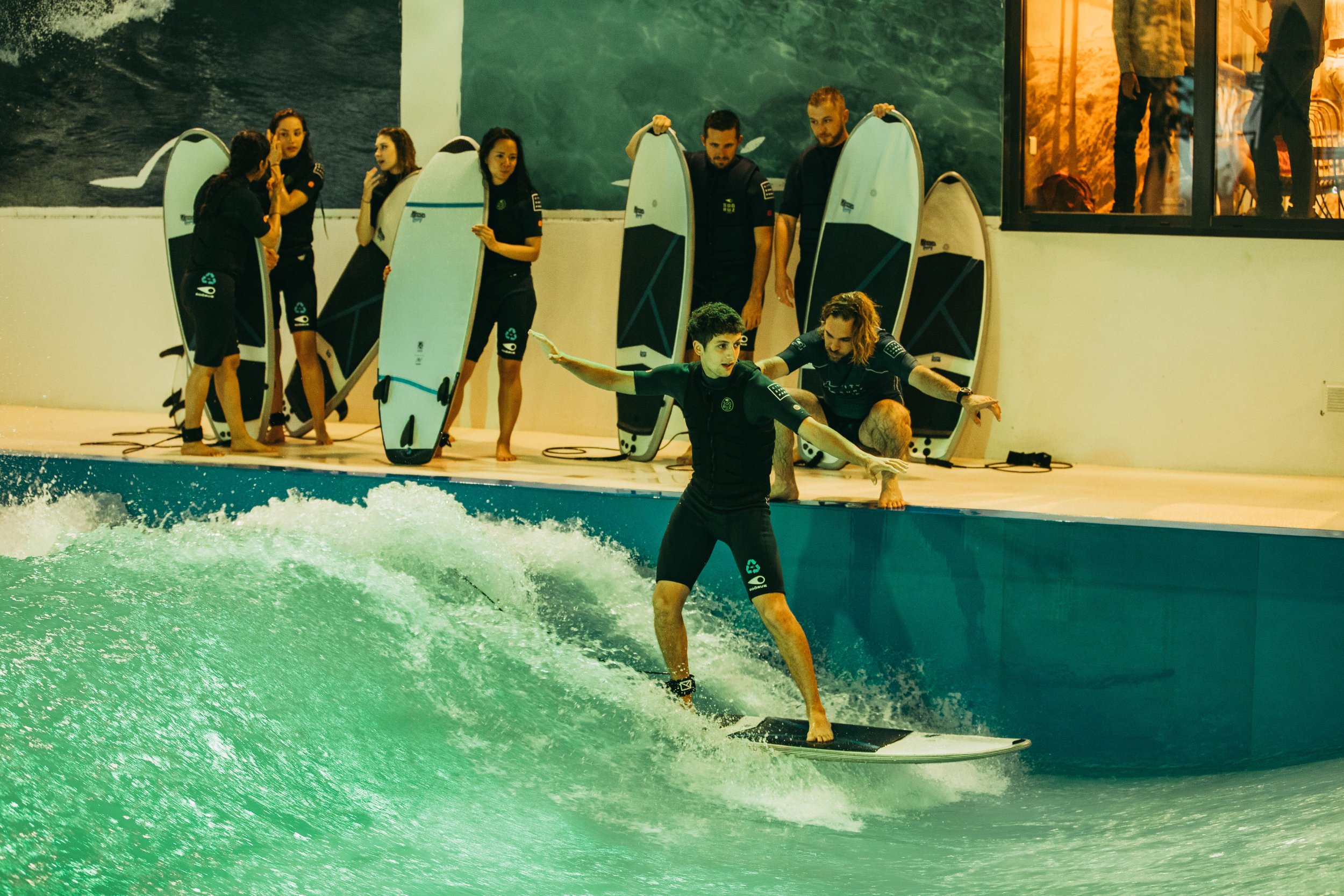 Personne pratiquant le surf en indoor, entourée d'équipe de support et de planches de surf, dans une piscine artificielle.