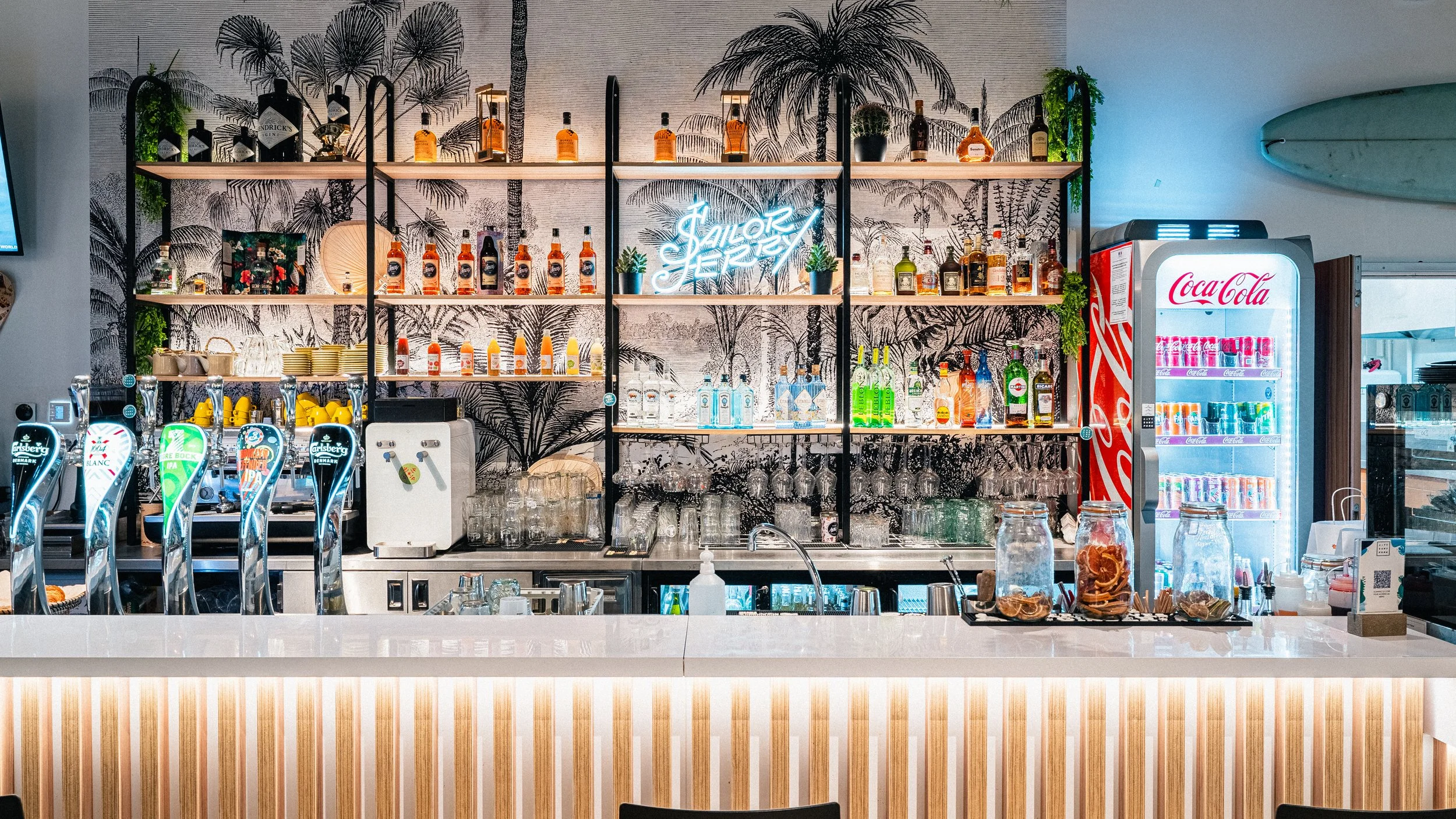 Bar avec étagères en métal et bois contenant des bouteilles d'alcool, avec un mur en papier peint à motifs de palmiers en arrière-plan, un neon lumineux "J'adore Jersey" au centre, et un frigo Coca-Cola lumineux à droite.