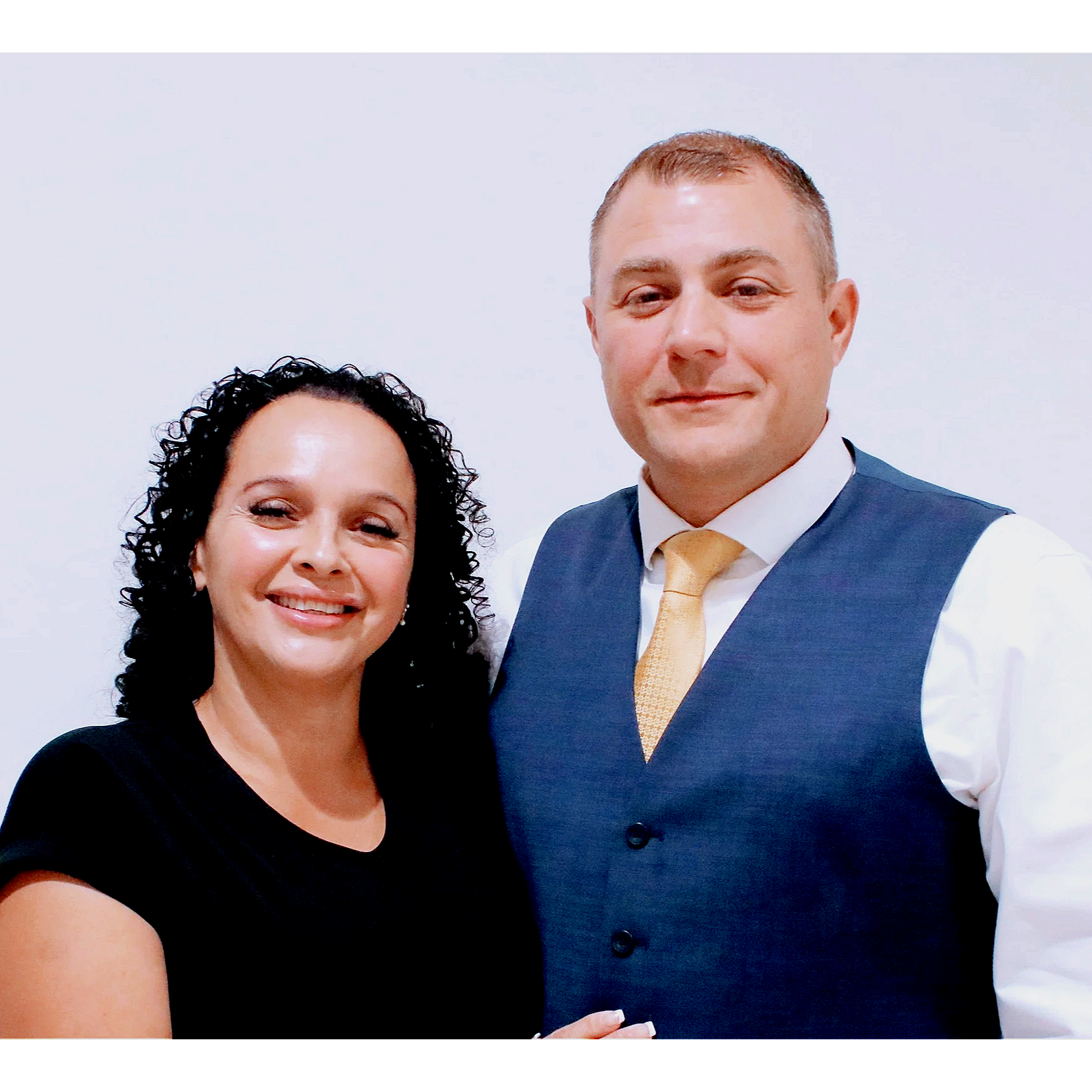 A smiling woman with dark, curly hair standing next to a man in a suit, white shirt, and gold tie, against a plain white background.