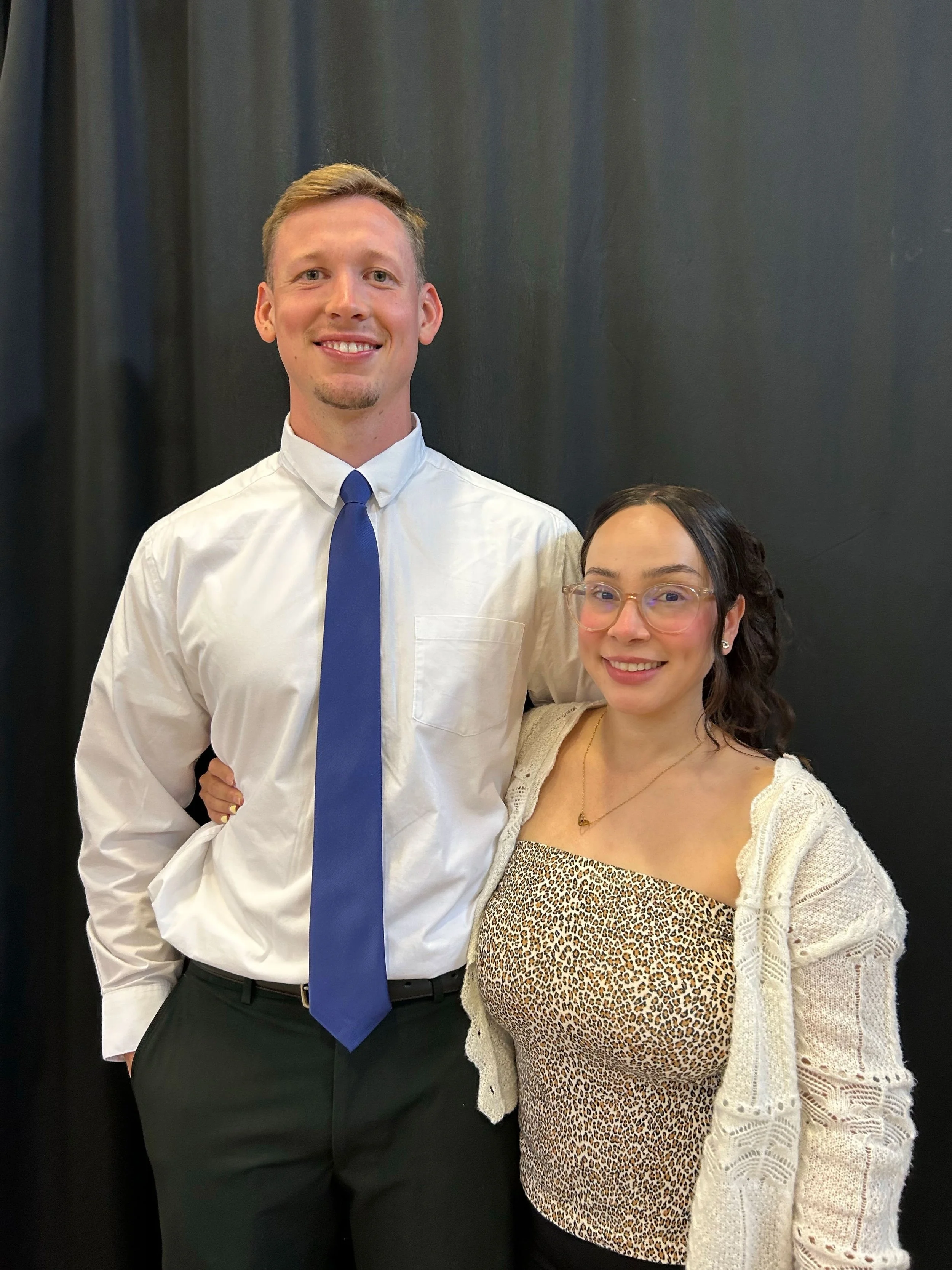 A man and a woman posing together in front of a dark curtain. The man wears a white shirt, blue tie, and black pants, and the woman wears glasses, a leopard print top, and a beige crochet cardigan.