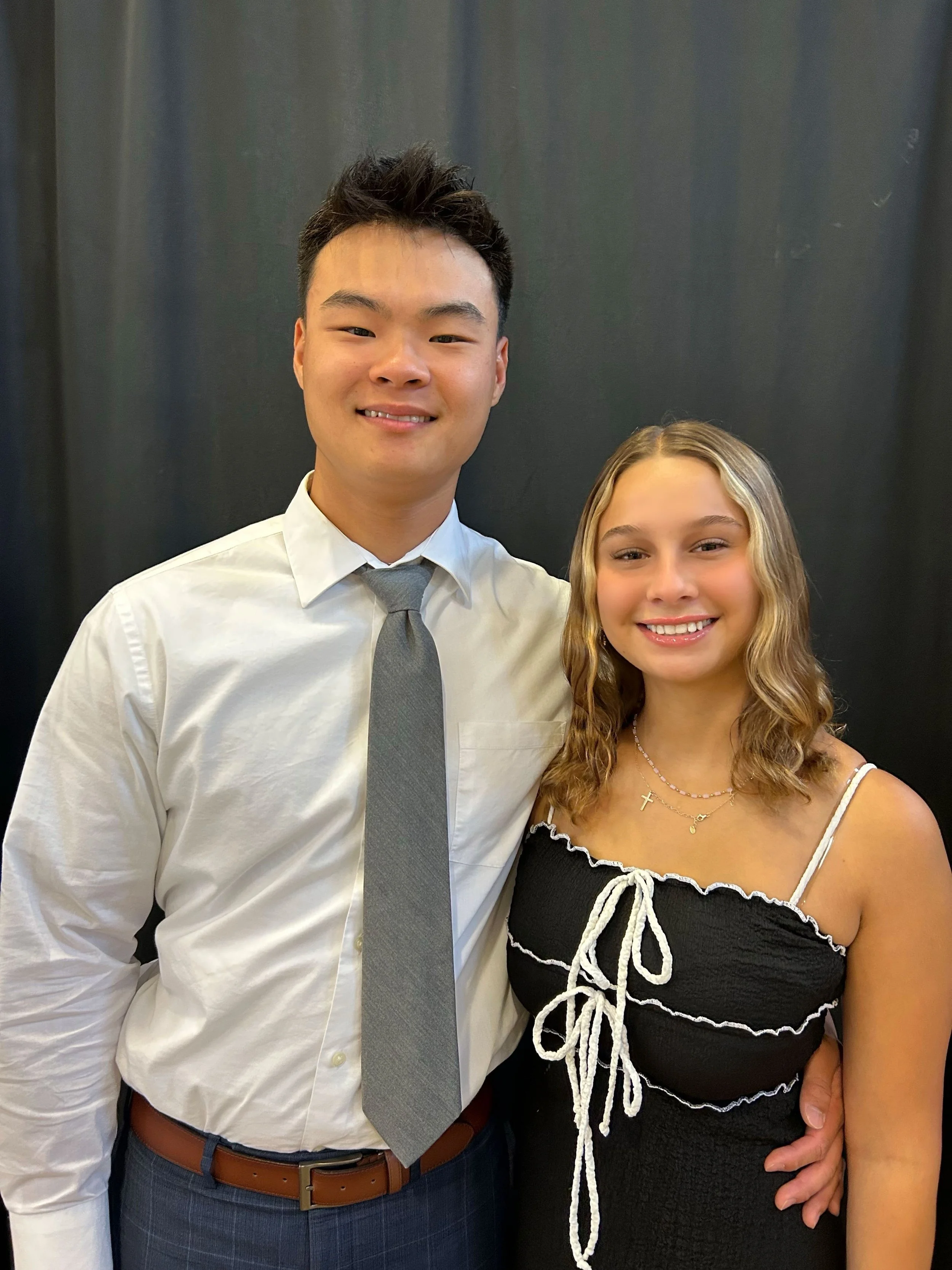 A man and a woman standing close together, smiling for a photo against a dark background. The man is wearing a white shirt, gray tie, and gray plaid pants. The woman is wearing a black top with white lace detailing and multiple necklaces.