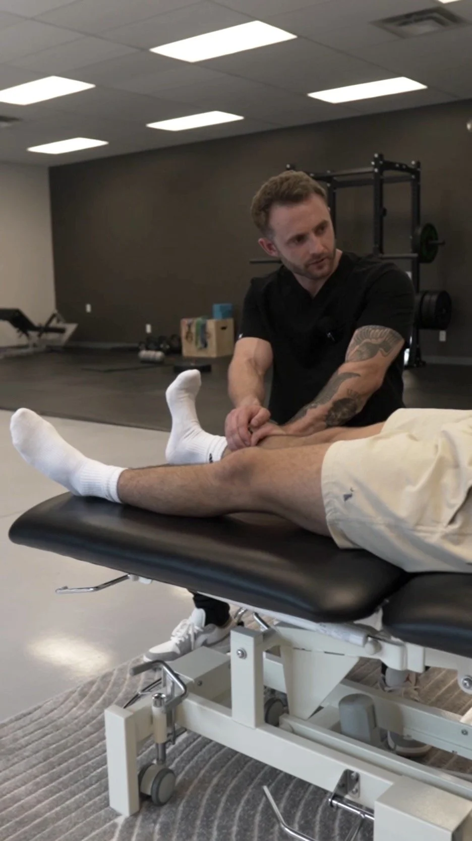 Physical therapist examining a patient's leg on an examination table in a gym or therapy room with workout equipment in the background.
