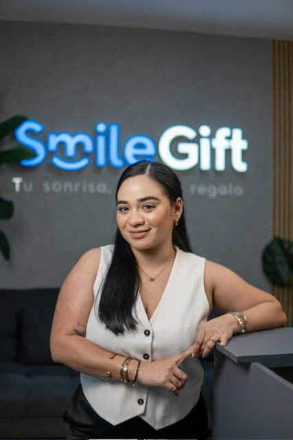 Mujer en la recepción de Smile Gift, una tienda de regalos, sonriendo y posando con fondo de letrero iluminado que dice 'Smile Gift' y el lema 'Tu sonrisa, nuestro regalo'.