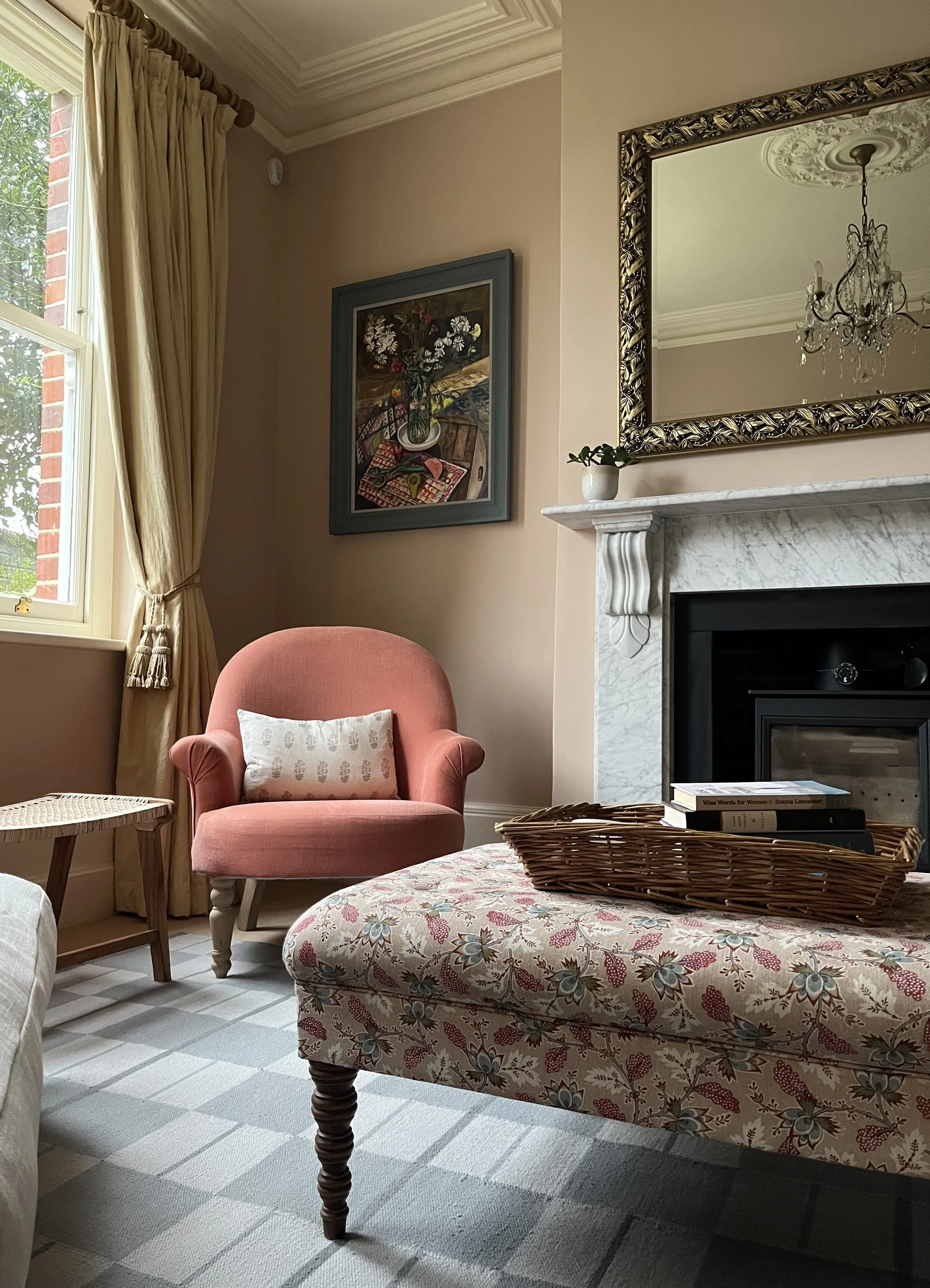 A cozy living room corner with a pink upholstered armchair, white and patterned ottoman, a marble fireplace with a gold-framed mirror above, a small potted plant on the mantel, and colorful framed artwork on the beige walls. Sunlight streams in through a window with beige curtains.