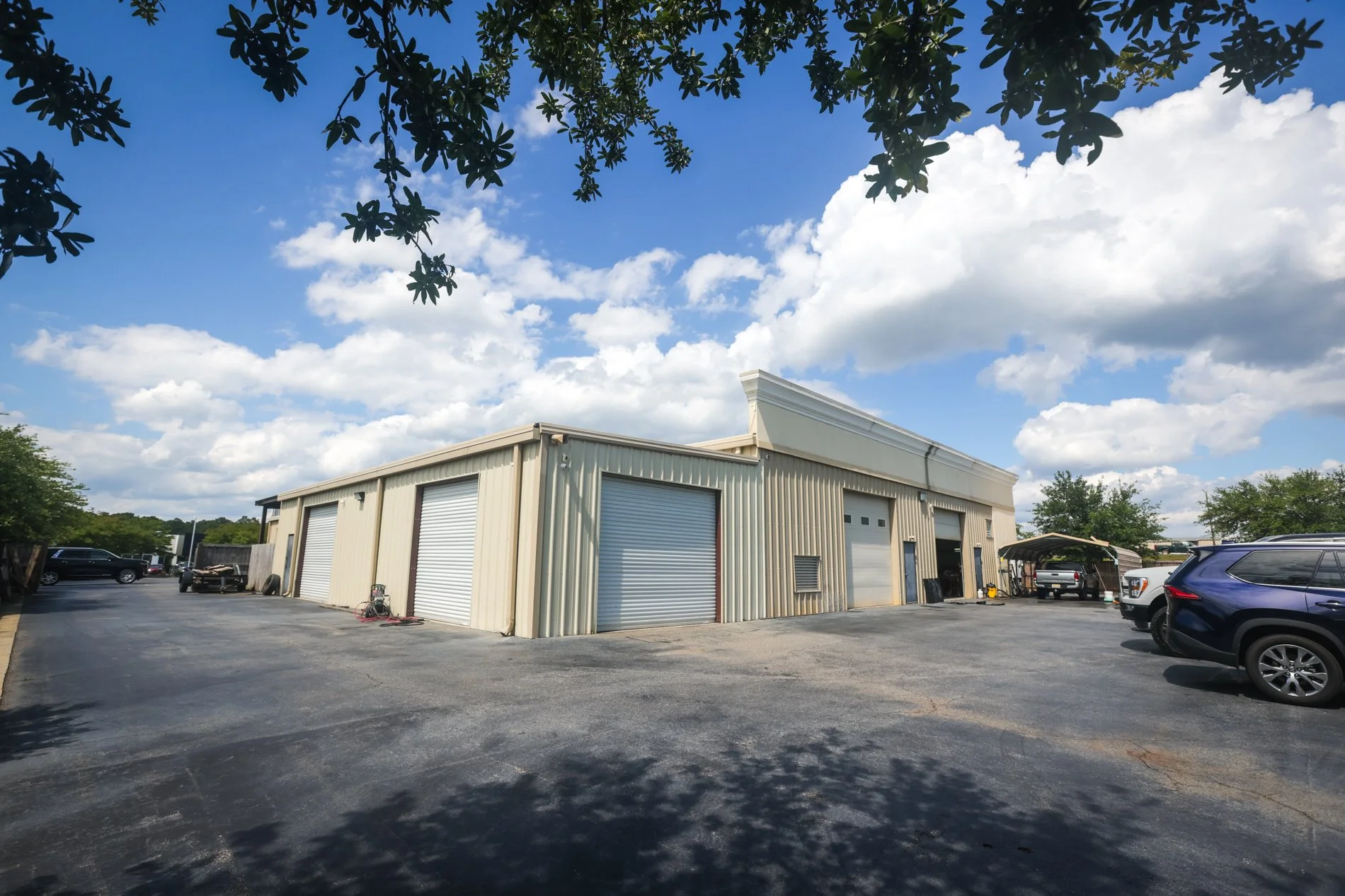 A metal building with multiple garage doors situated in a parking lot, with vehicles parked nearby and a partly cloudy sky overhead.