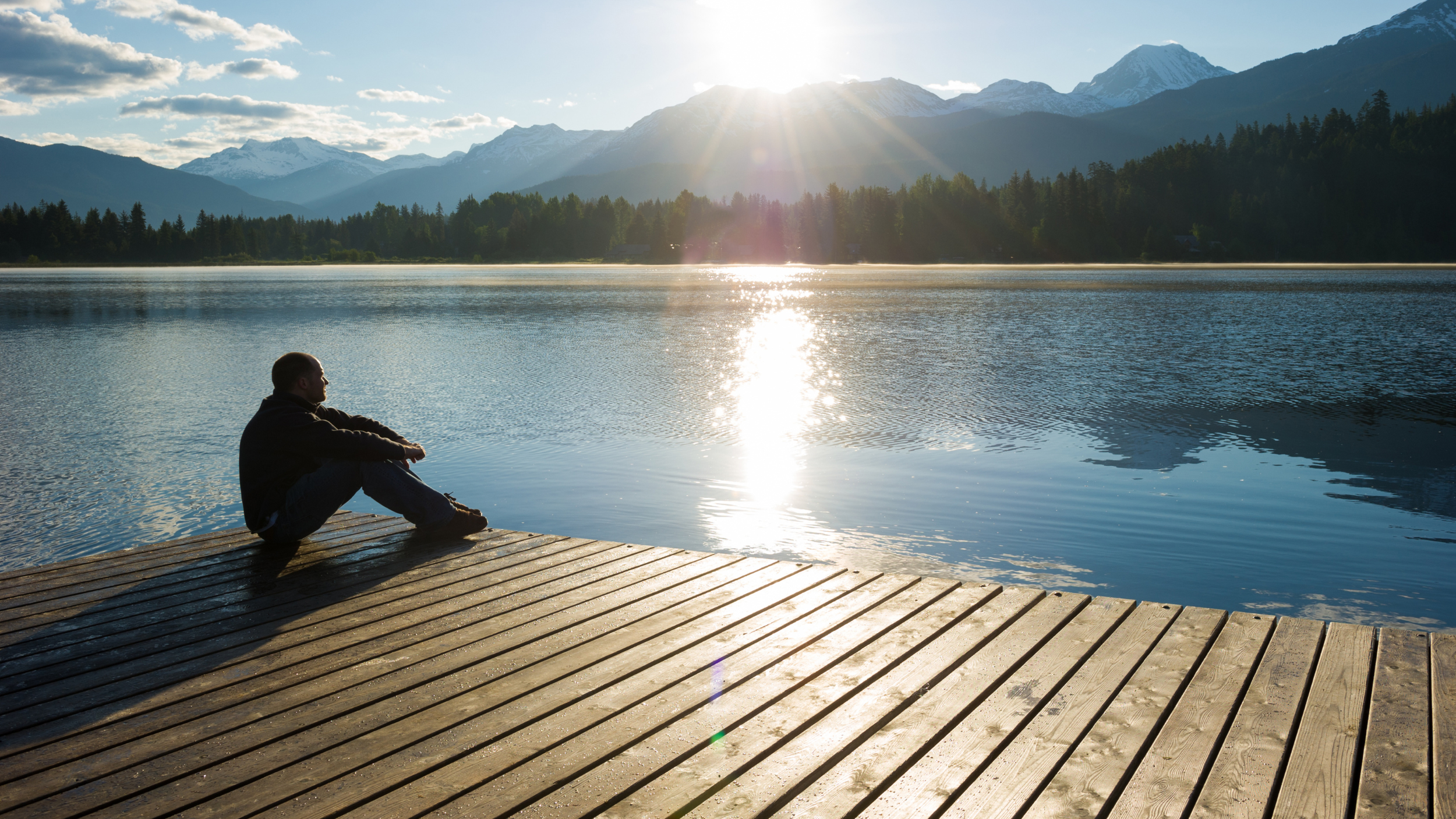 person sitting by lake and mountains looking sun's reflection