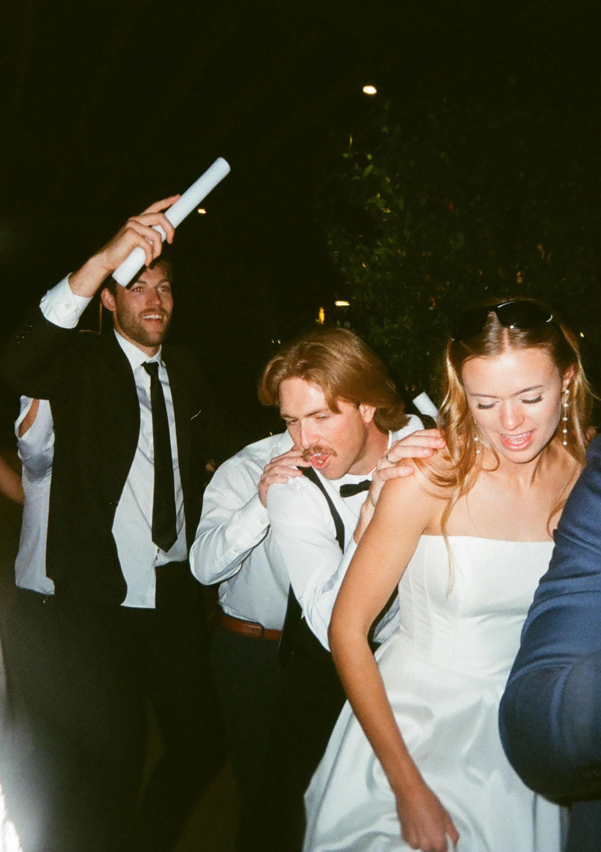 People dancing at a celebration or party, some wearing formal attire, dancing closely together, with a dark outdoor background.