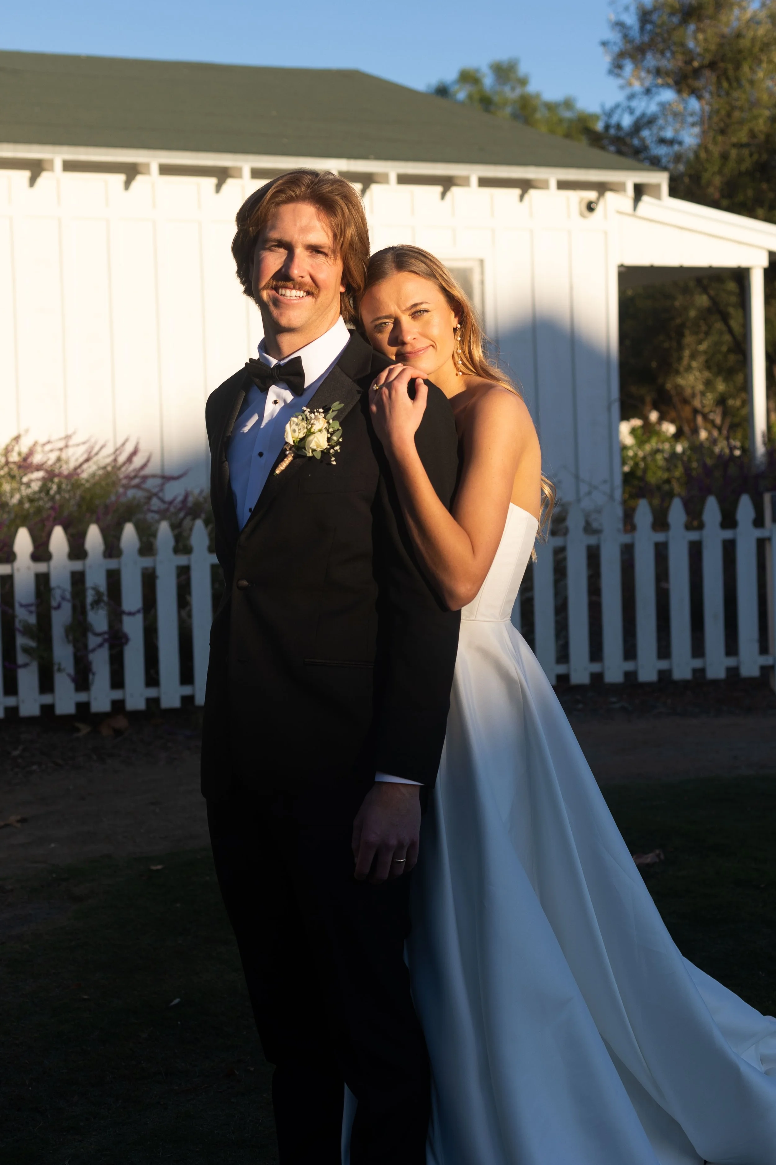 A newlywed couple on their wedding day standing outdoors in front of a white fence and house, with the bride hugging the groom from behind, both smiling.