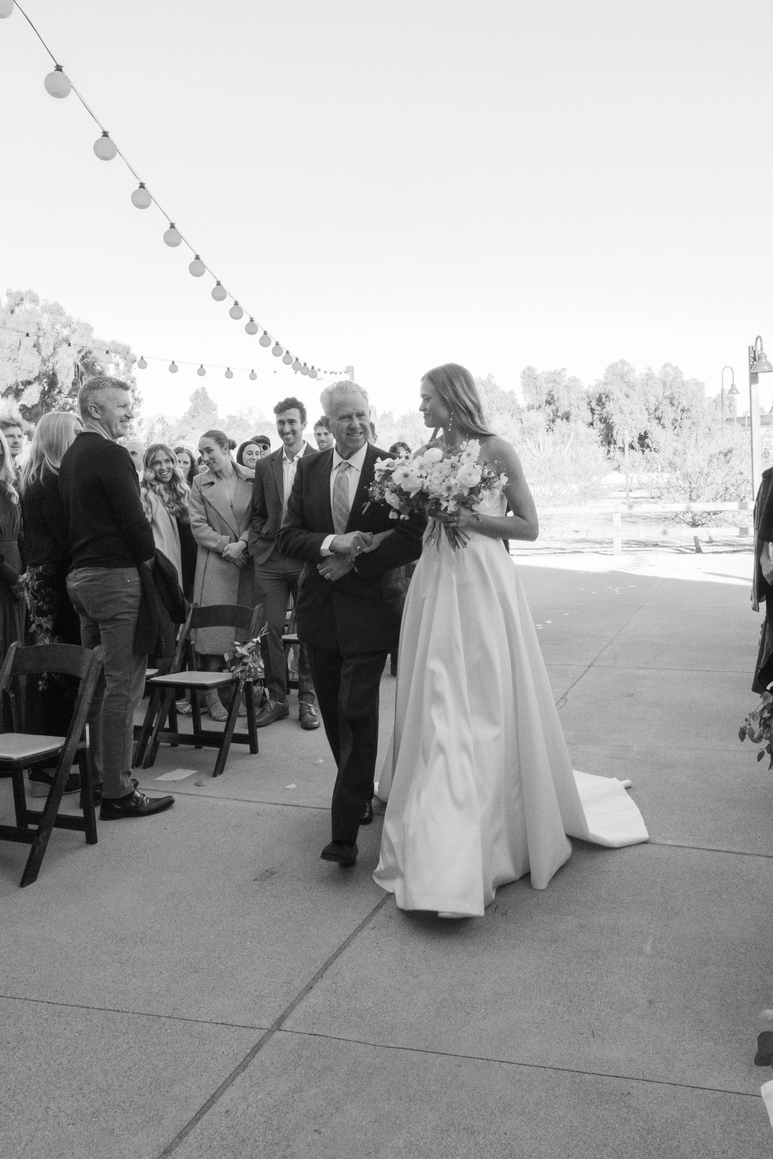 A bride in a wedding gown holding a bouquet of flowers walking down the aisle, being escorted by an older man, surrounded by wedding guests outdoors under string lights.