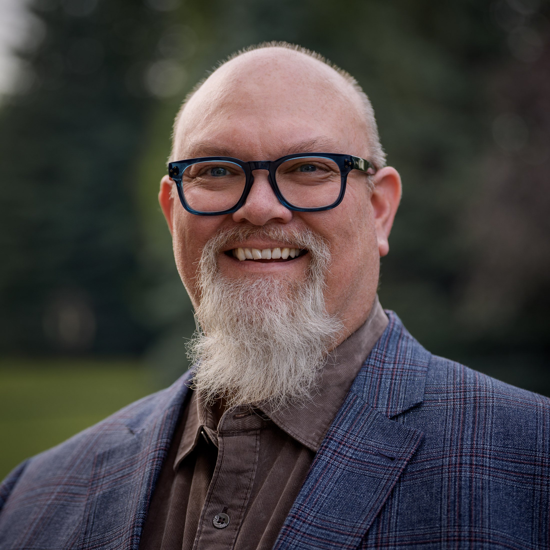 Close-up of a man with glasses, bald head, white goatee, and smiling, wearing a brown shirt and blazer outdoors.