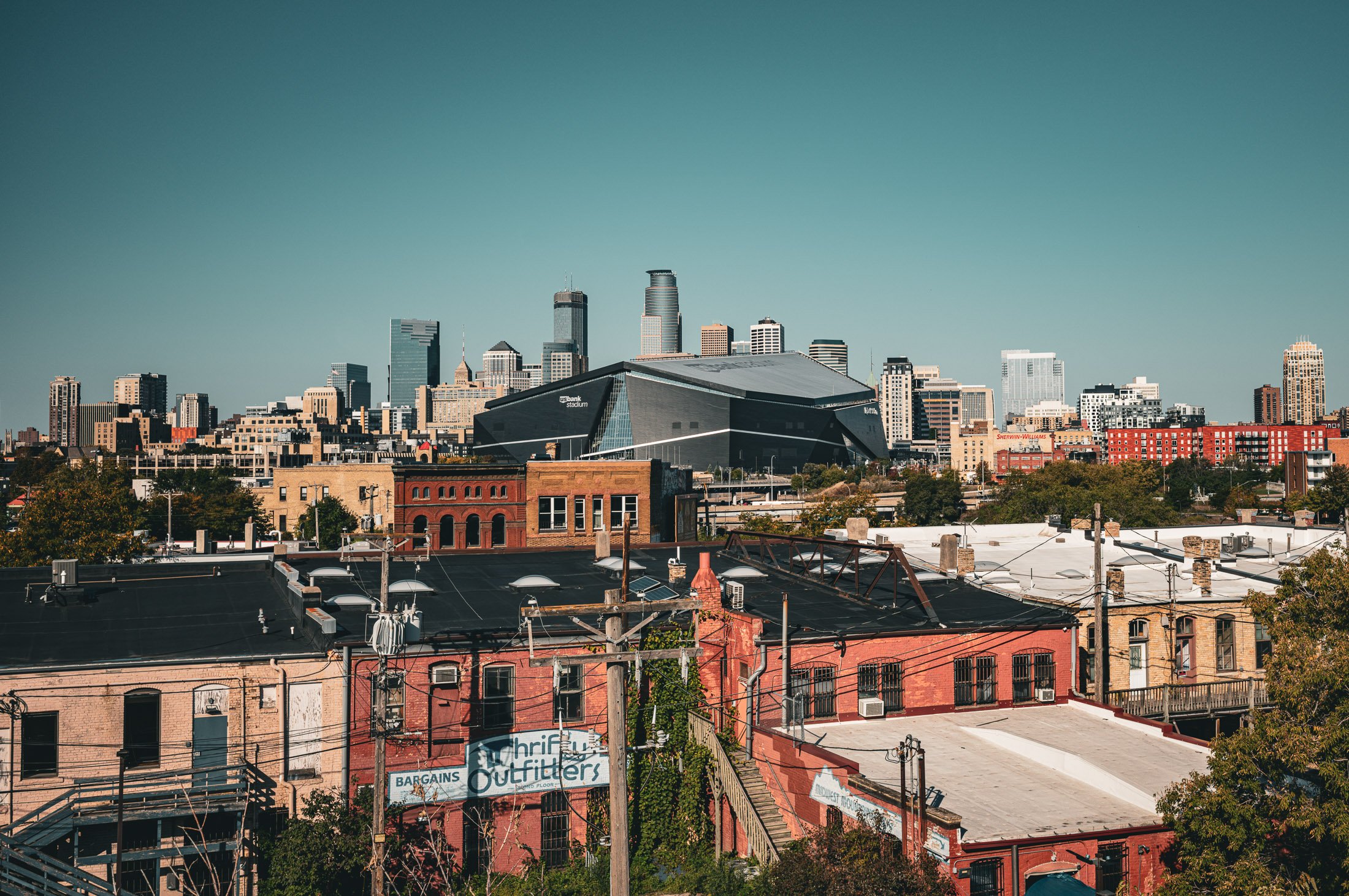 Minneapolis skyline with downtown skyscrapers in the distance and older brick buildings in the foreground, including the U.S. Bank stadium and billboards.