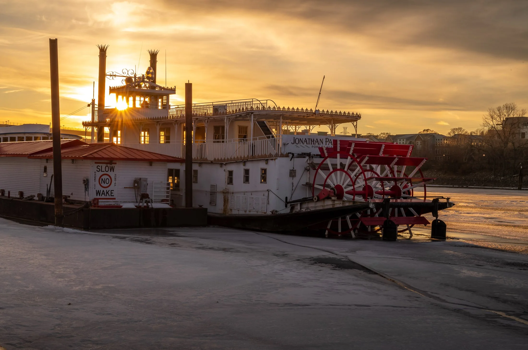 A historic paddlewheel riverboat docked at a harbor during sunset, with the sun partially obscured by the boat’s upper deck, creating a starburst effect. The boat has a white exterior with red accents, including a large red paddlewheel at the stern, 