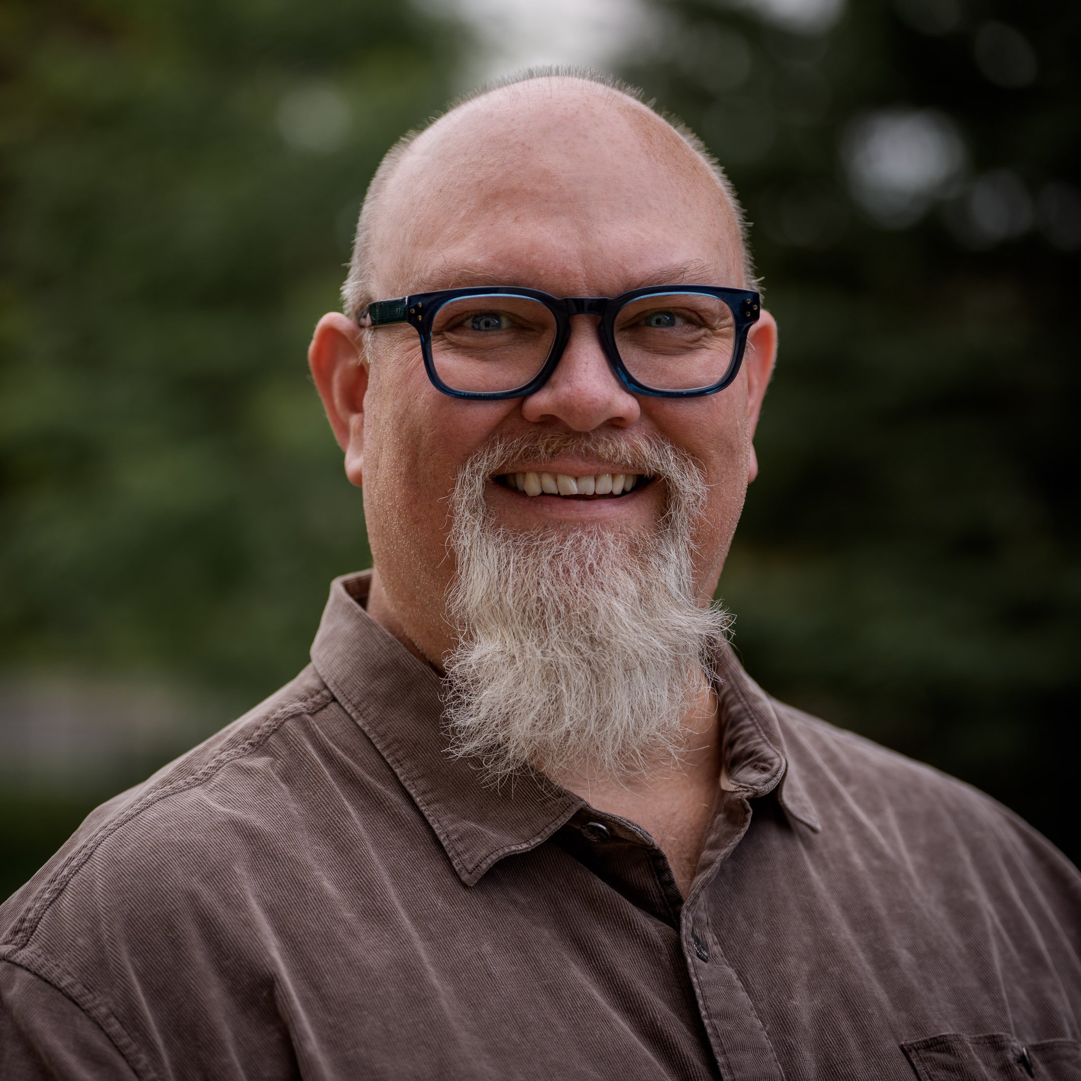 Close-up of a smiling man with a bald head and white goatee, wearing black-rimmed glasses, smiling outdoors against blurred greenery background.