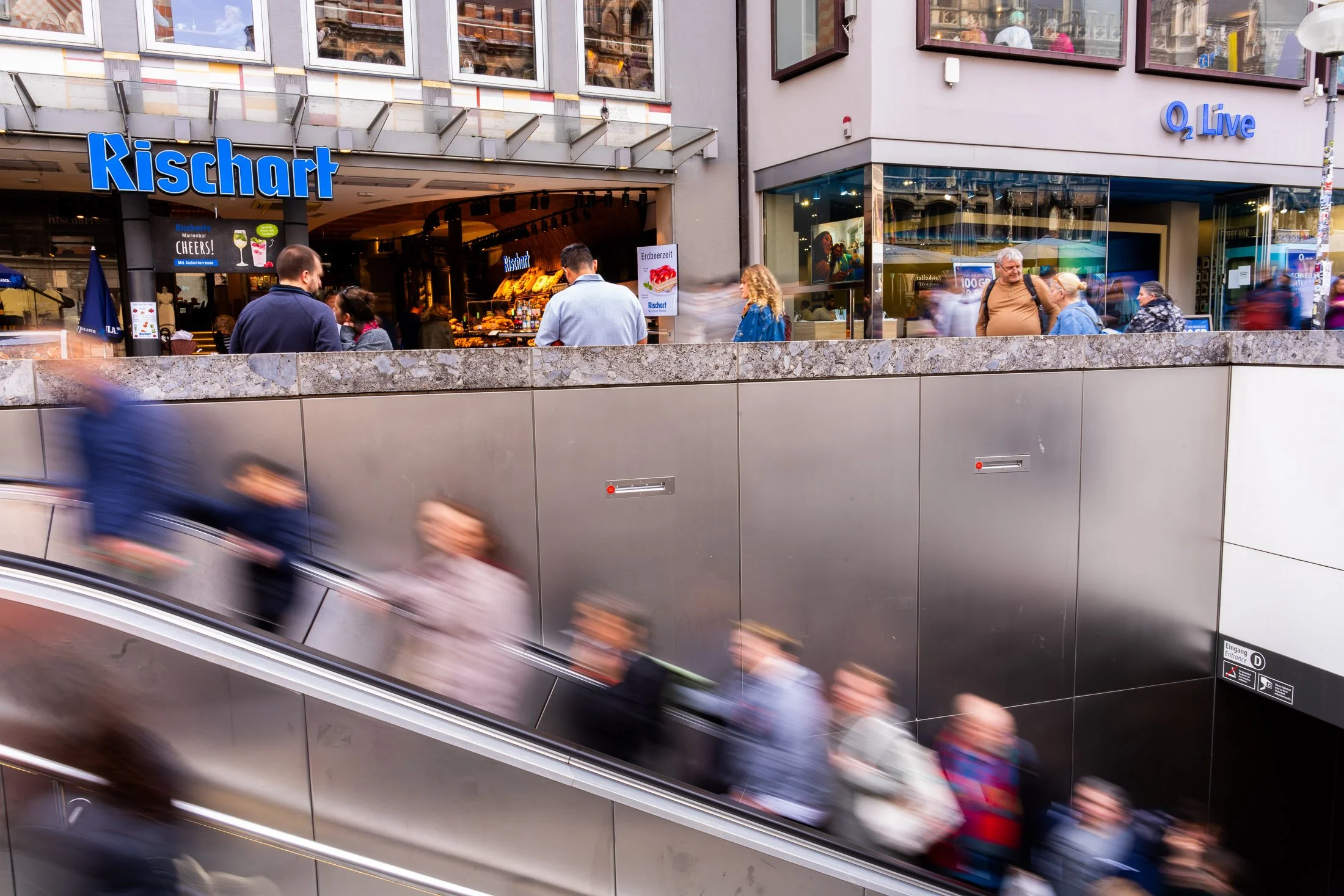 People in motion on an outdoor subway escalator in Germany near others who are walking and talking outside a store called Rischart and an O2 Live store, with some individuals visible through the glass windows on an urban street.