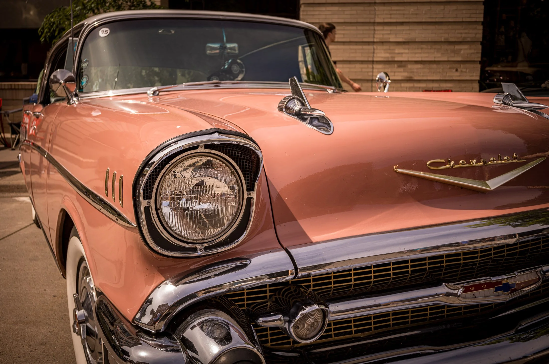 Close-up of a vintage pink Chevrolet car with chrome details and custom side mirrors, parked outdoors with buildings in the background.