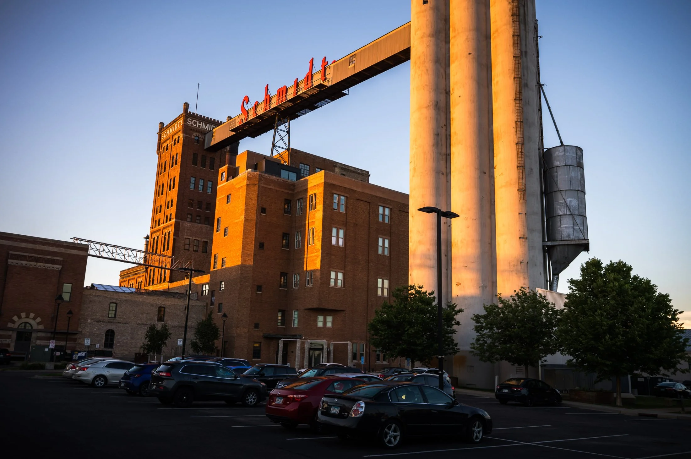 An urban scene featuring a parking lot filled with cars in front of a brick building. Above the building, there is a large sign that reads 'Schmidt,' supported by a metal structure extending from the building. The scene is illuminated by warm sunligh