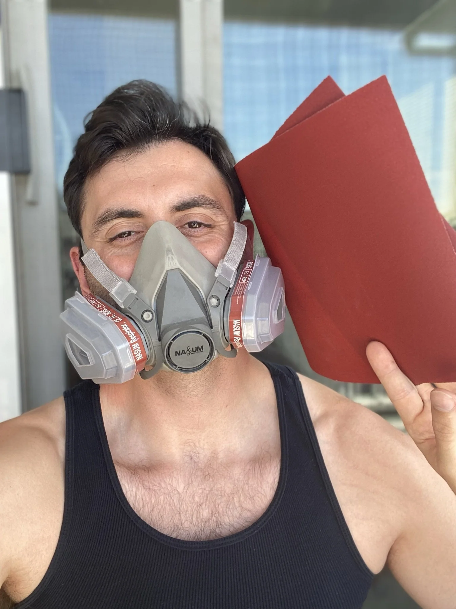 A man wearing a protective mask is holding a red folder or piece of sandpaper, smiling outside.