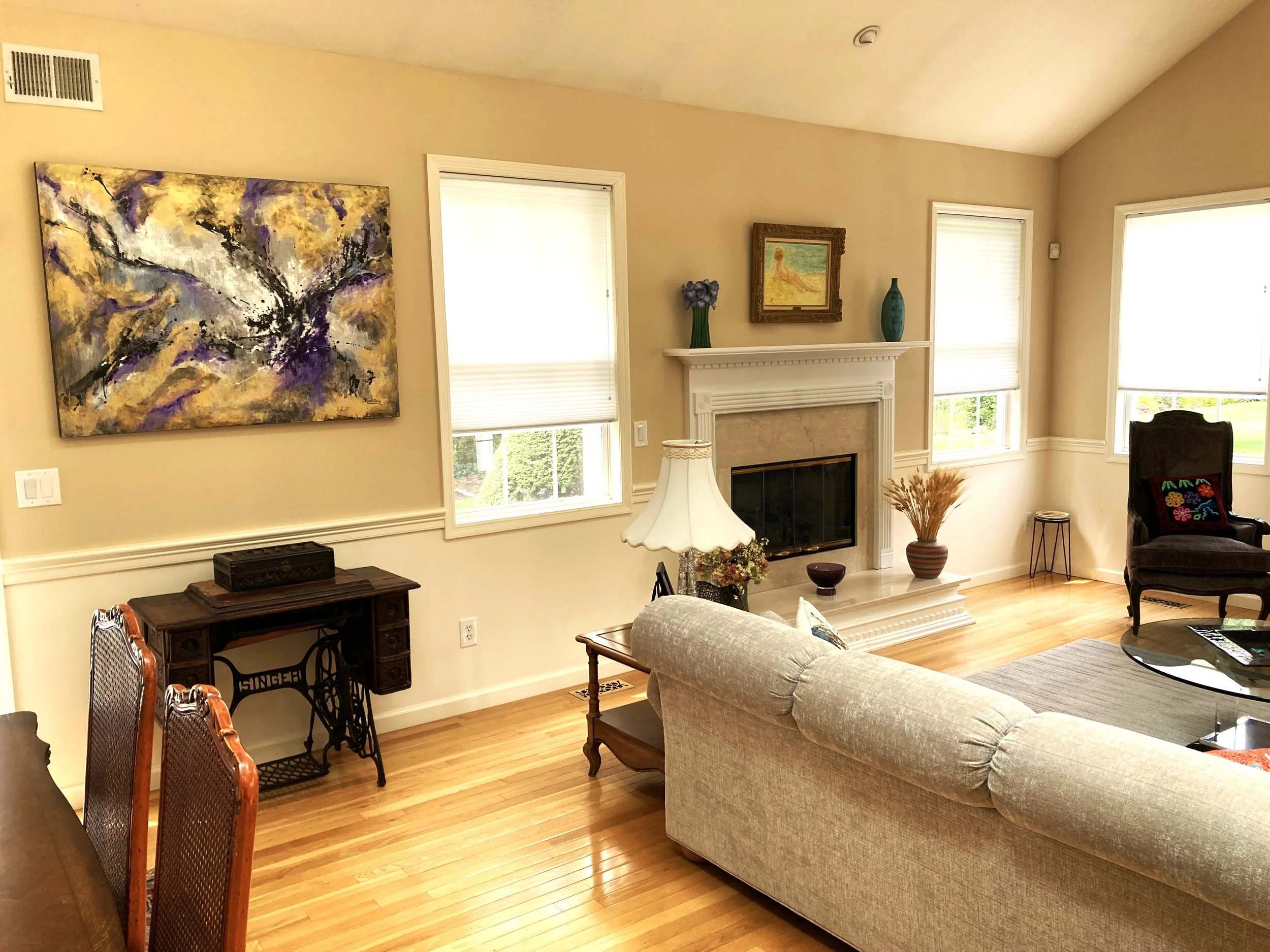 Living room with beige walls, hardwood floor, fireplace with mantel, beige sofa, black armchair, wooden side table with lamp, wall art, and windows with blinds that let in natural light.