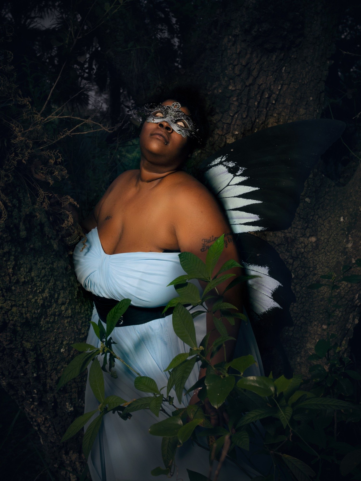 A woman with dark skin lying against a tree trunk, wearing a silver masquerade mask, a white dress with black accents, large black and white fairy wings, and surrounded by green leaves in a natural, wooded environment.