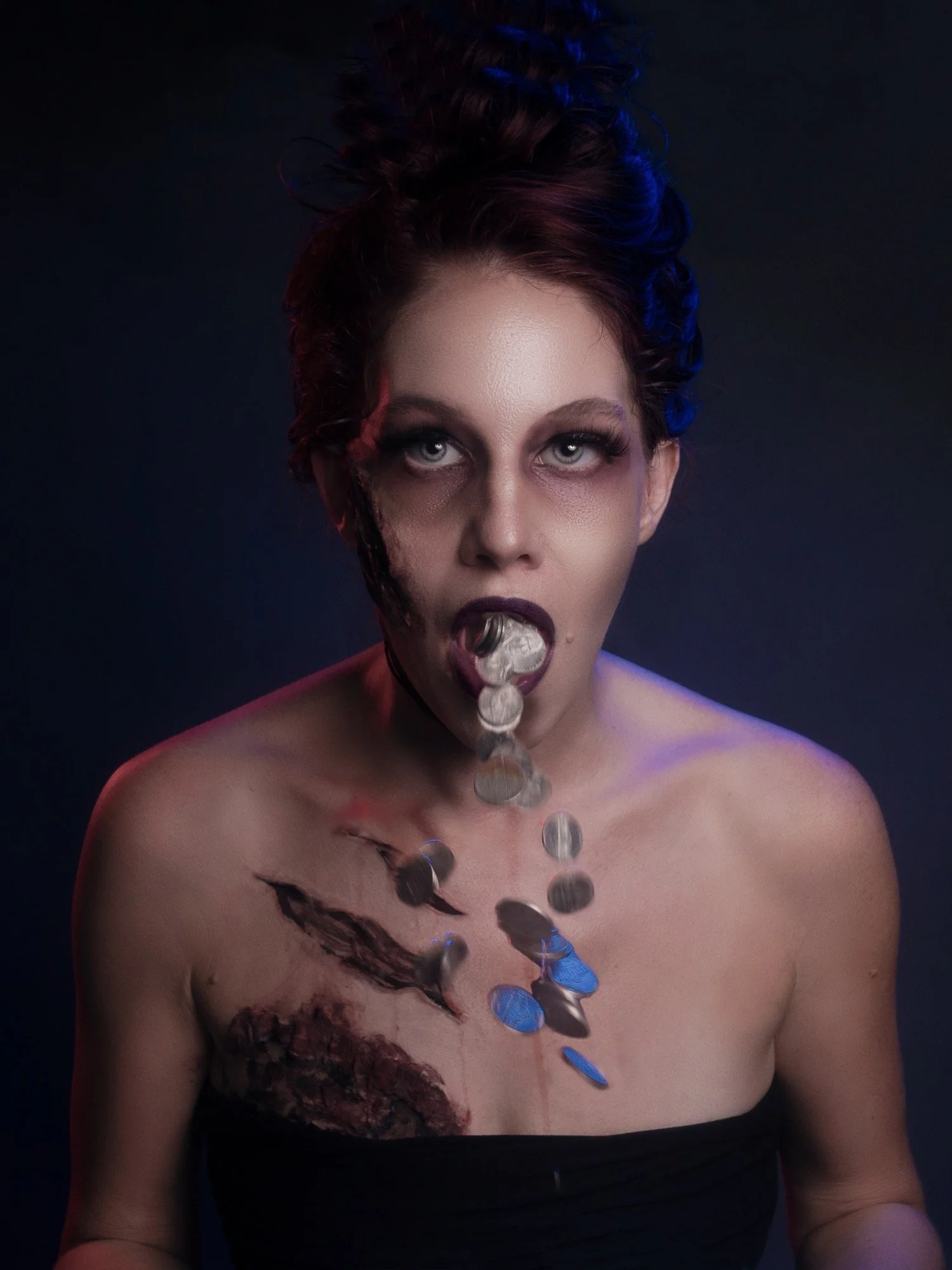 A woman with dark hair styled in a messy bun, dark makeup, and tattoos on her shoulder and chest, is sticking her tongue out, catching falling silver coins against a dark background.