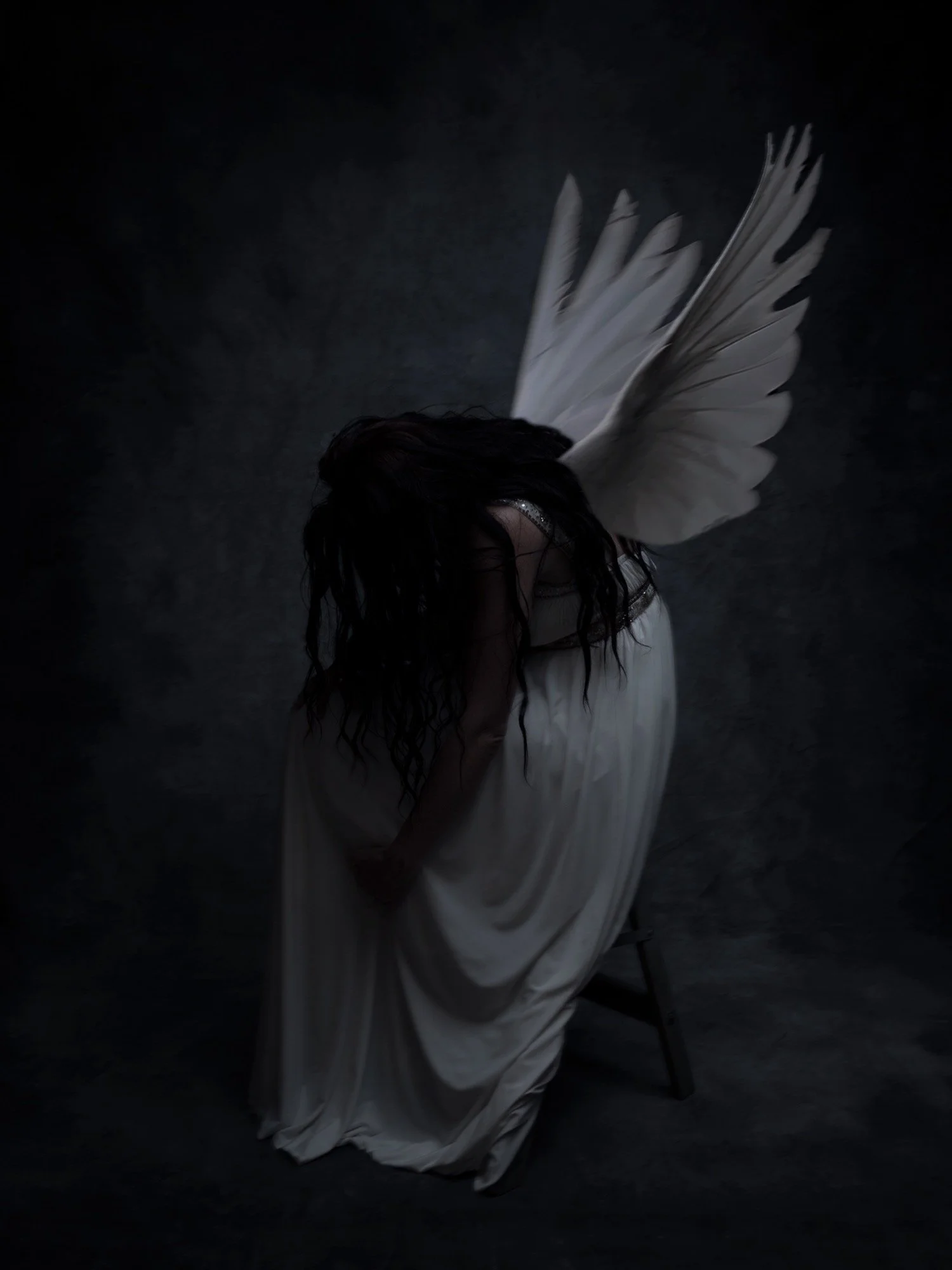 A person dressed as an angel with large white wings and a flowing white gown, sitting on a small stool, with dark hair covering their face, in a dimly lit setting.
