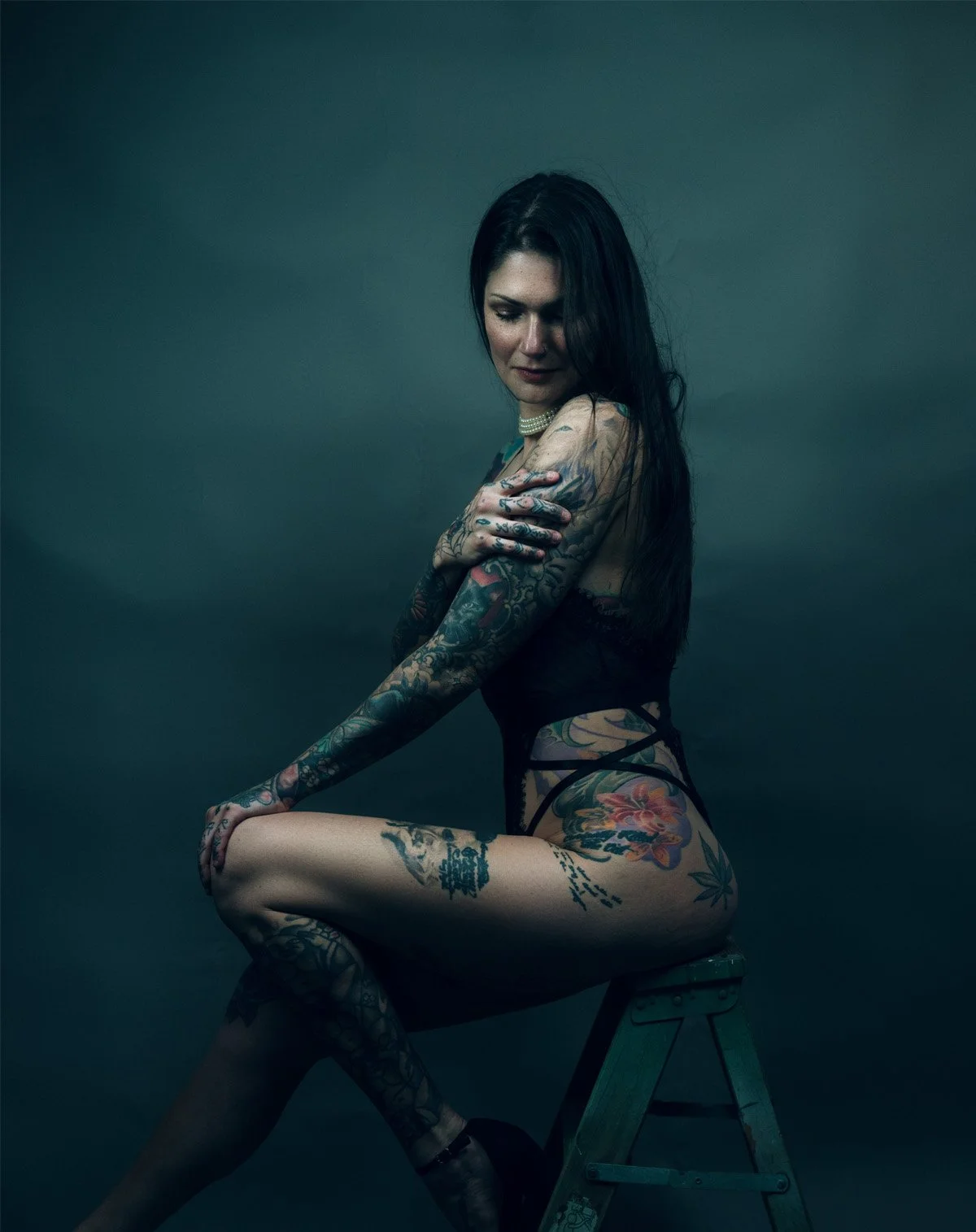A tattooed woman with long dark hair and pearl necklace sitting on a wooden stool against a dark background, wearing black lingerie.