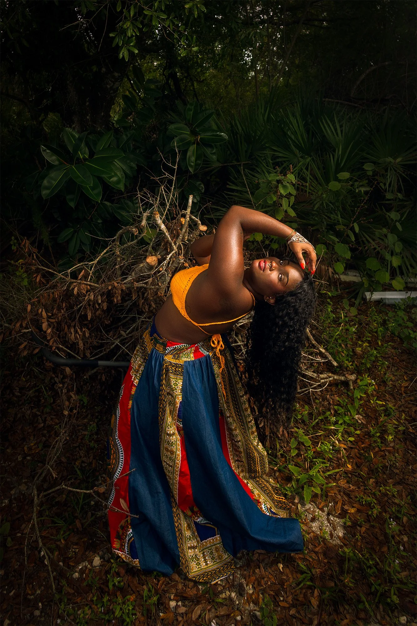 A woman with dark skin and long curly hair is posing outdoors among green plants and fallen leaves, wearing colorful traditional clothing, with one arm raised behind her head and the other resting on her face.