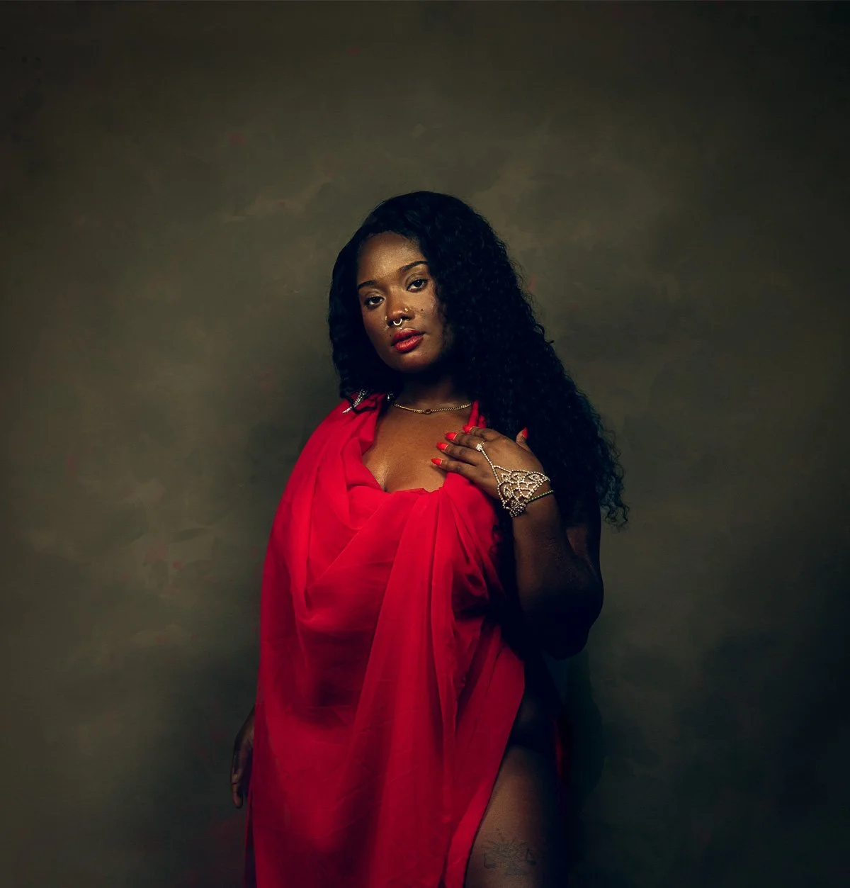 A woman with long, curly black hair wearing a red dress and jewelry, posing against a dark background.