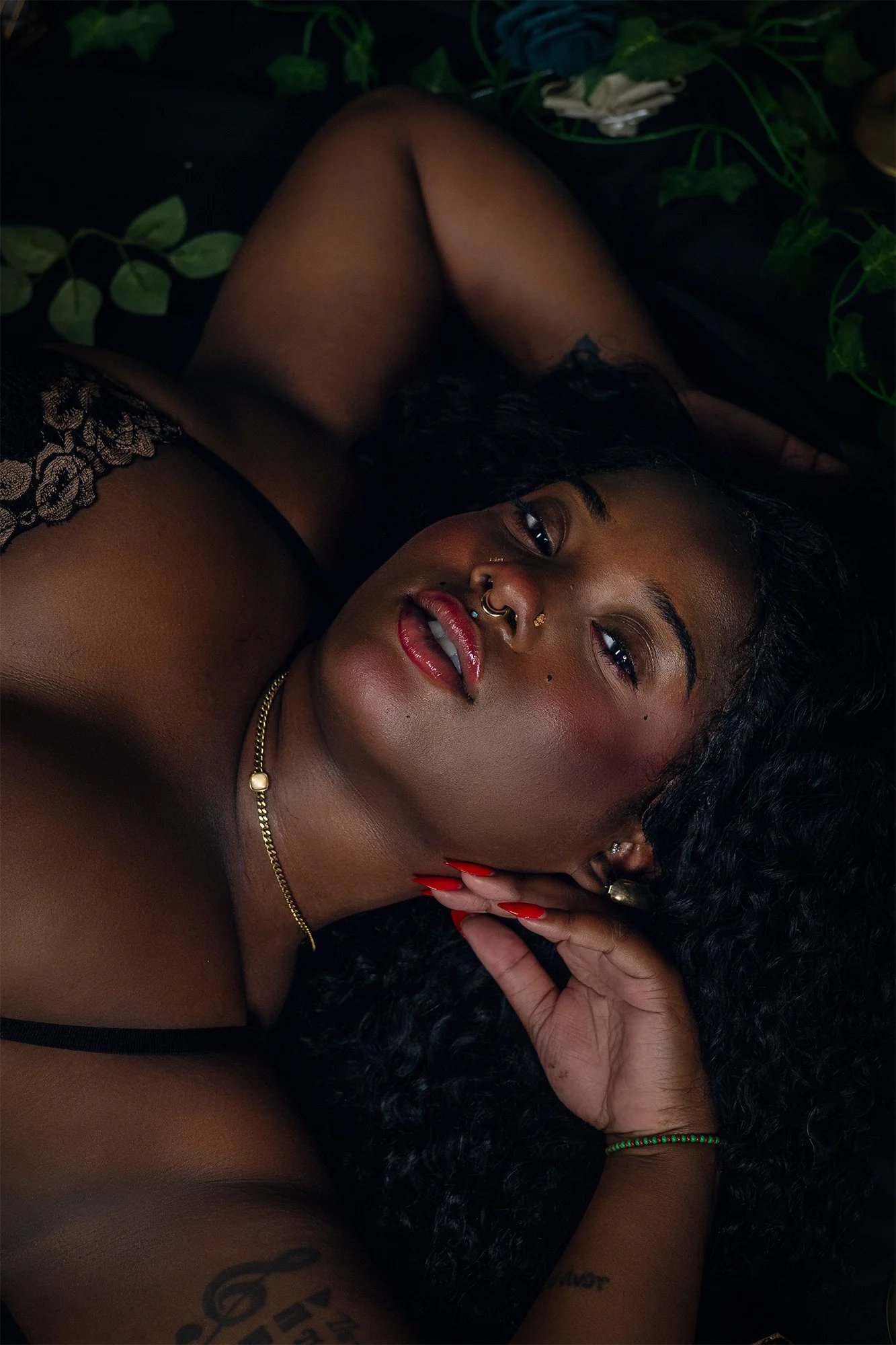 A woman with curly black hair lying down surrounded by green leaves, wearing lace black lingerie, red nail polish, and jewelry, looking at the camera.
